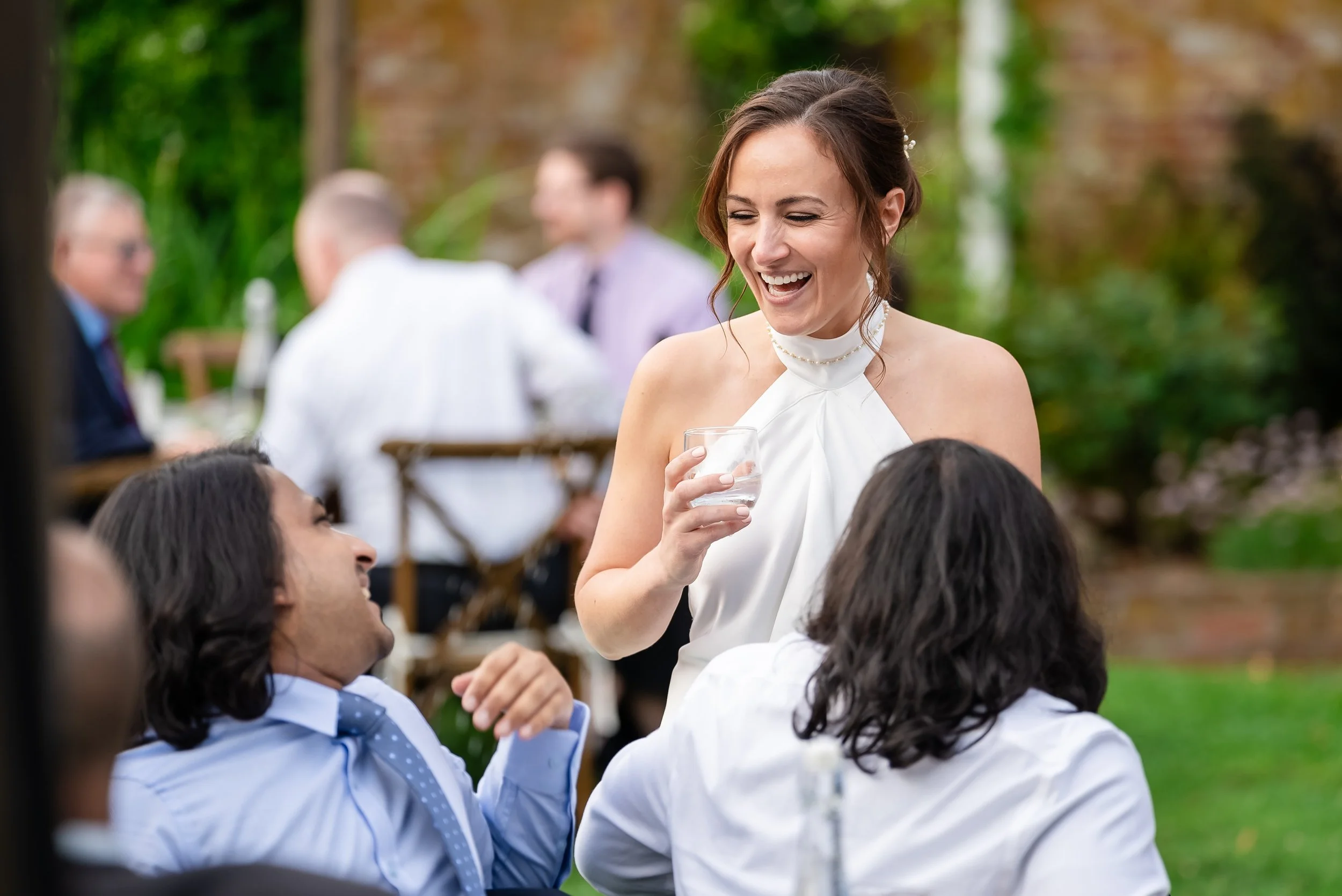 A woman in a white dress and pearl necklace laughing while talking to a man in a blue shirt and tie at an outdoor gathering.