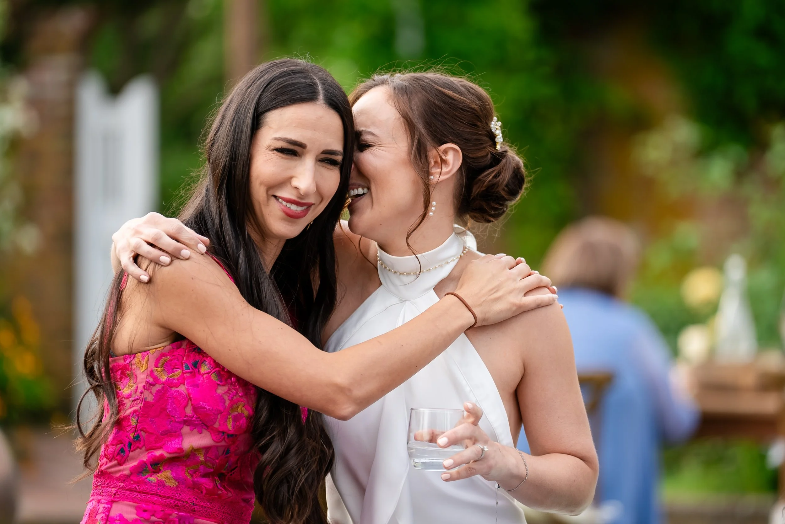 Two women hugging at an outdoor event, one with dark hair wearing a pink dress and the other with brown hair wearing a white dress, holding a glass of water.