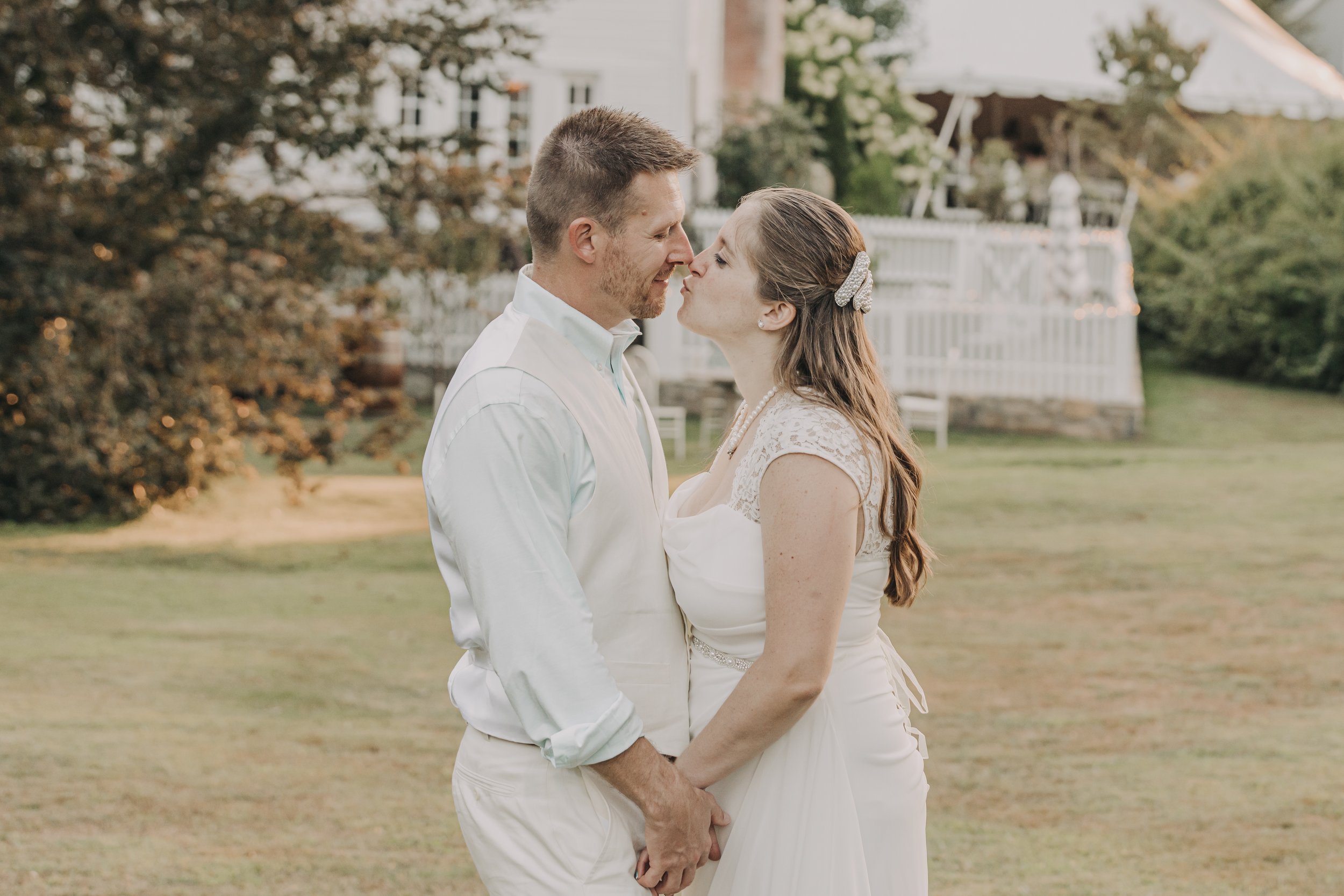 A man and woman stand close together outdoors during sunset, with their foreheads touching and smiling at each other. The woman wears a white dress with lace details and pearls, and a decorative hair clip. The man wears a white shirt, and they are ho