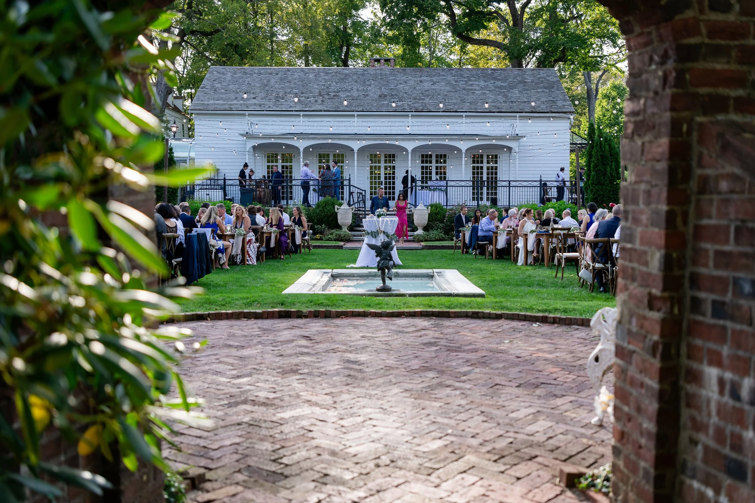 Outdoor gathering with tables and guests in front of a white house decorated with string lights, seen through a brick and greenery archway.