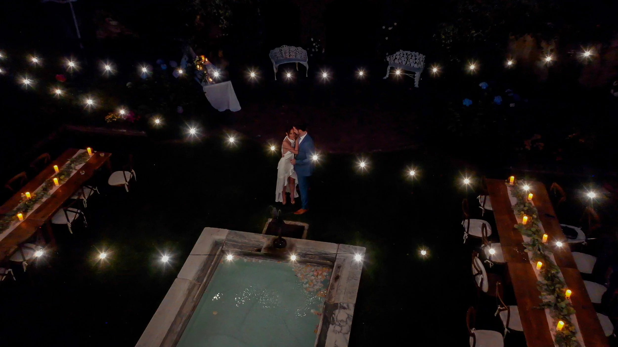 A couple dancing closely in the center of a dimly lit outdoor wedding reception area, decorated with candles and string lights, with tables and chairs surrounding them.