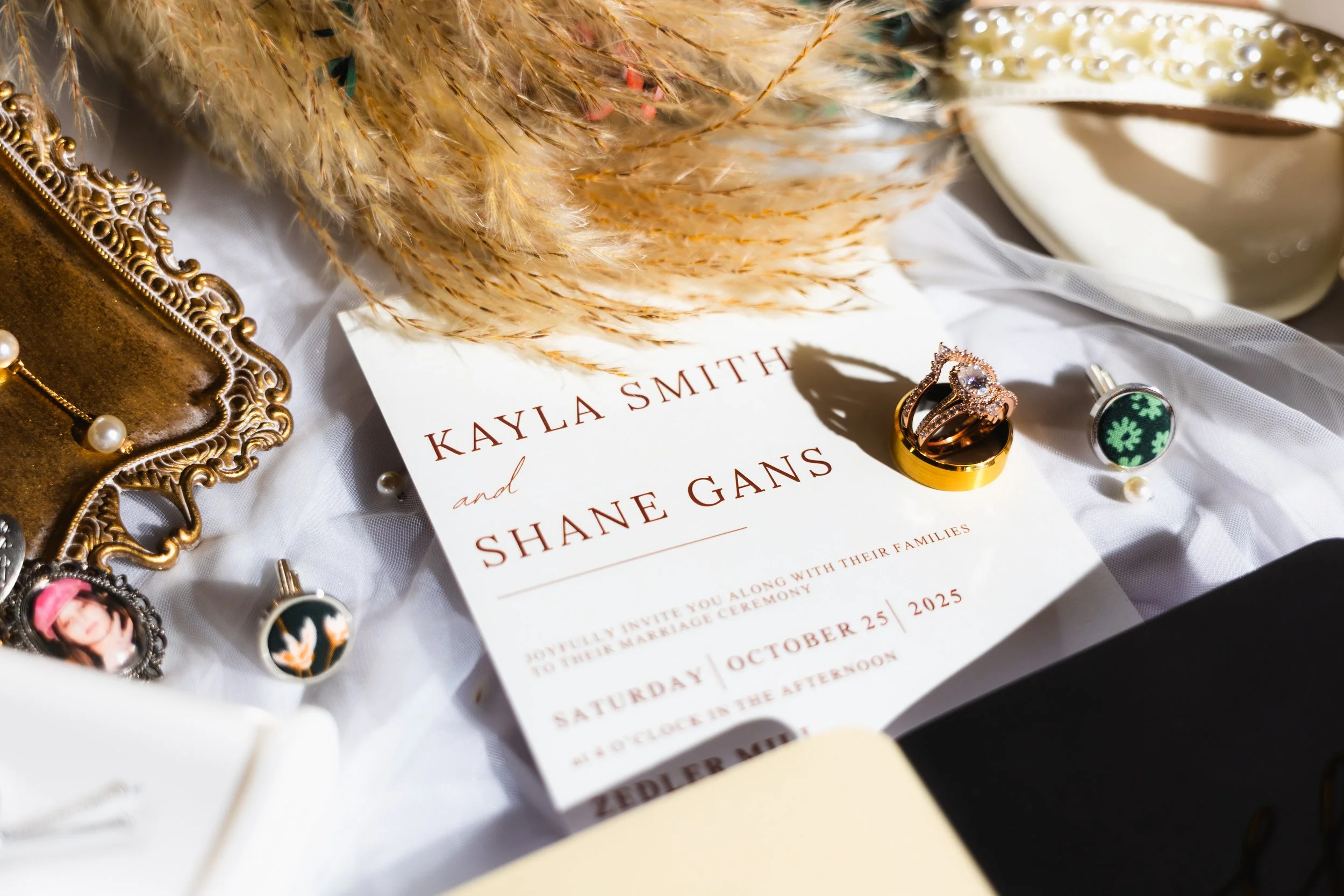 Wedding invitation card with gold rings, earrings, and jewelry on a white cloth, surrounded by dried flowers and other decorative items.