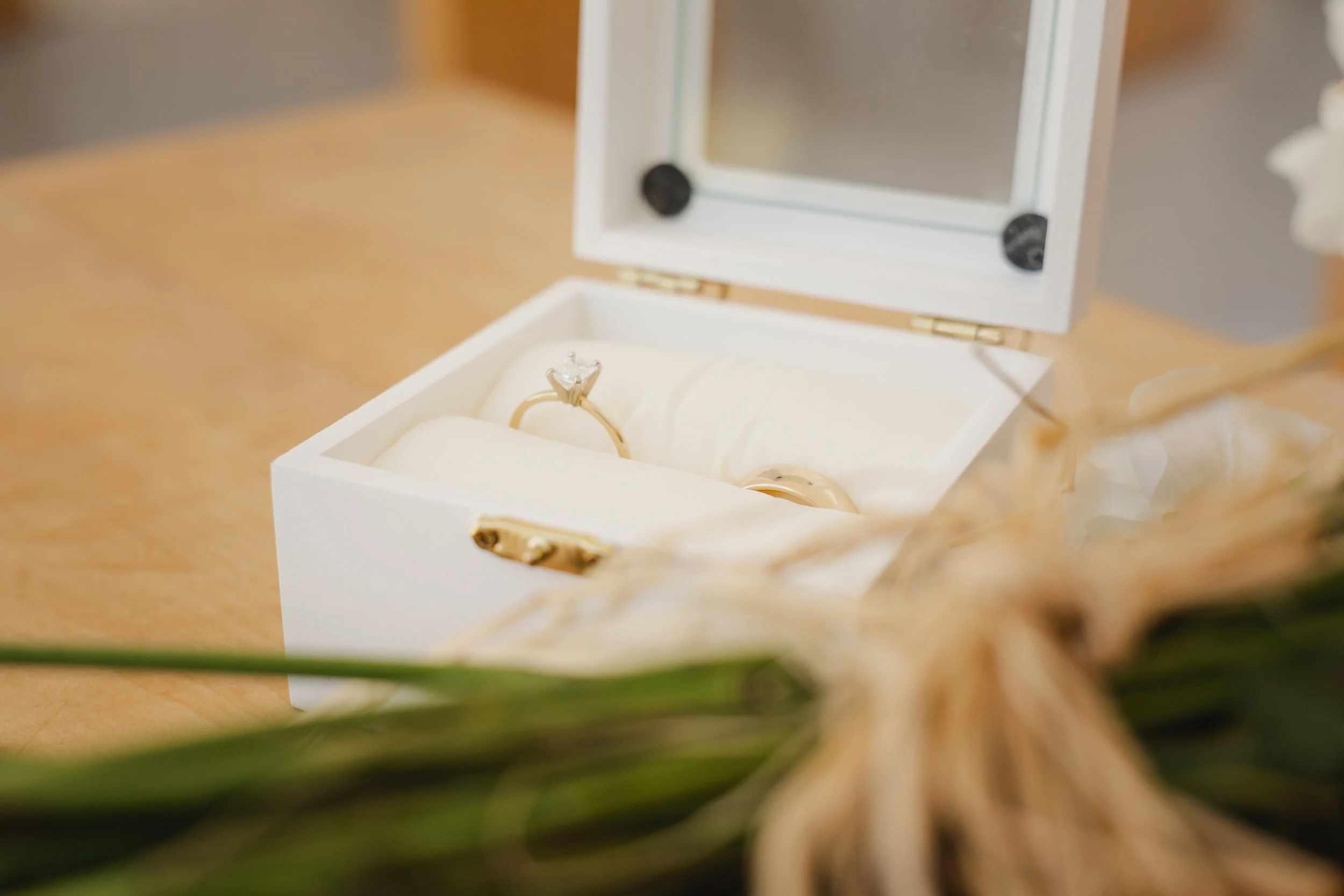 Open white jewelry box containing a diamond engagement ring and a wedding band, situated on a wooden surface with a blurred bouquet in front.
