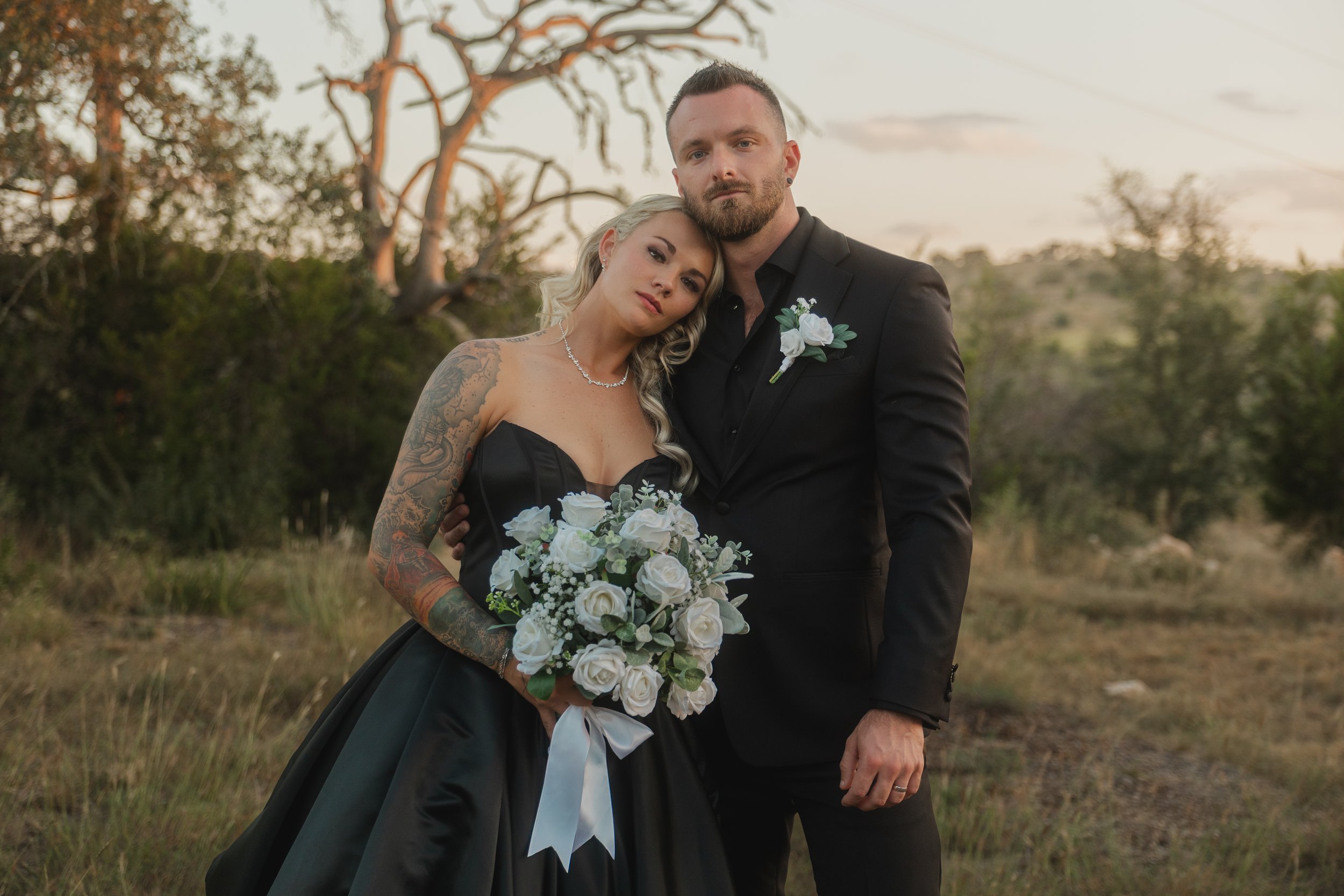 A bride and groom are standing outdoors during sunset, with the bride holding a bouquet of white roses and wearing a black dress, and the groom wearing a black suit with a white boutonniere, in a natural setting with trees and a grassy landscape in t
