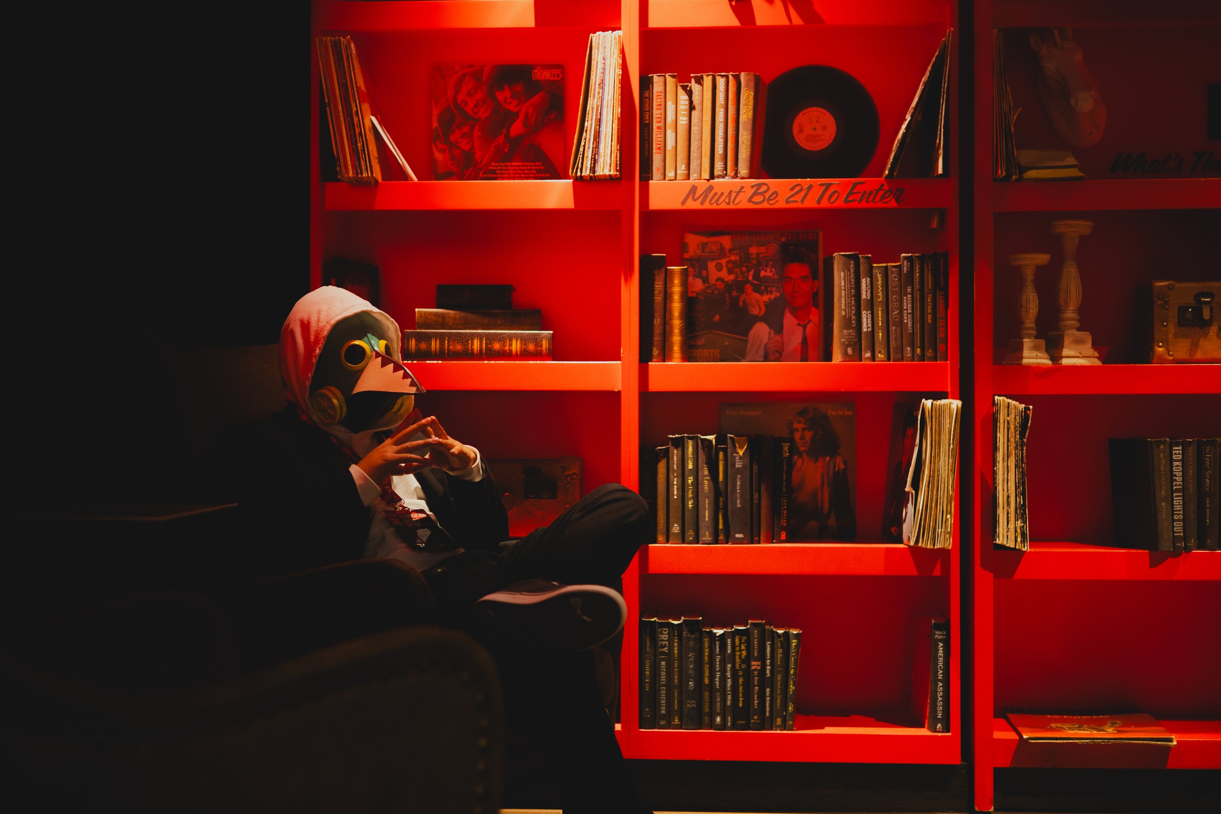 Person wearing a mask and headphones sitting on a black chair in front of a red bookshelf filled with vinyl records and books, dimly lit.