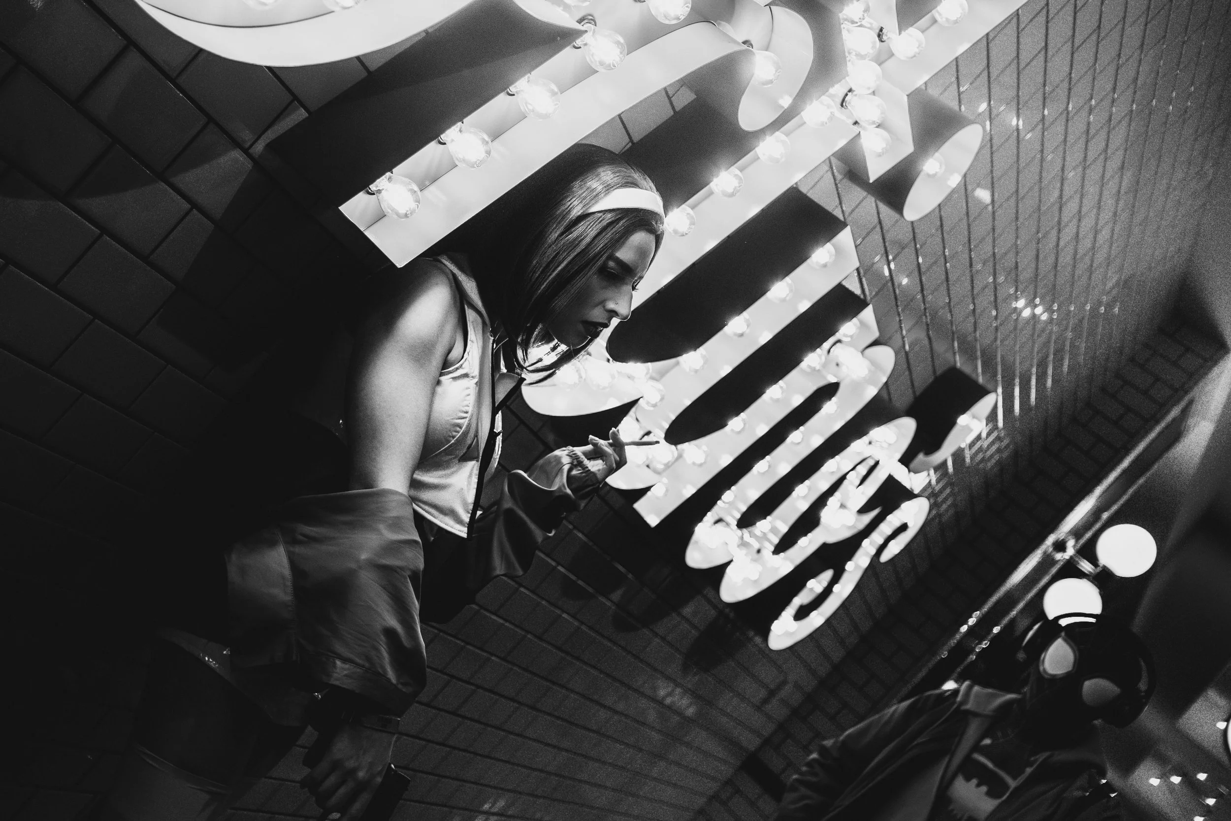 A woman standing against a brick wall with illuminated signs and decorative lights, looking down and holding a cigarette.