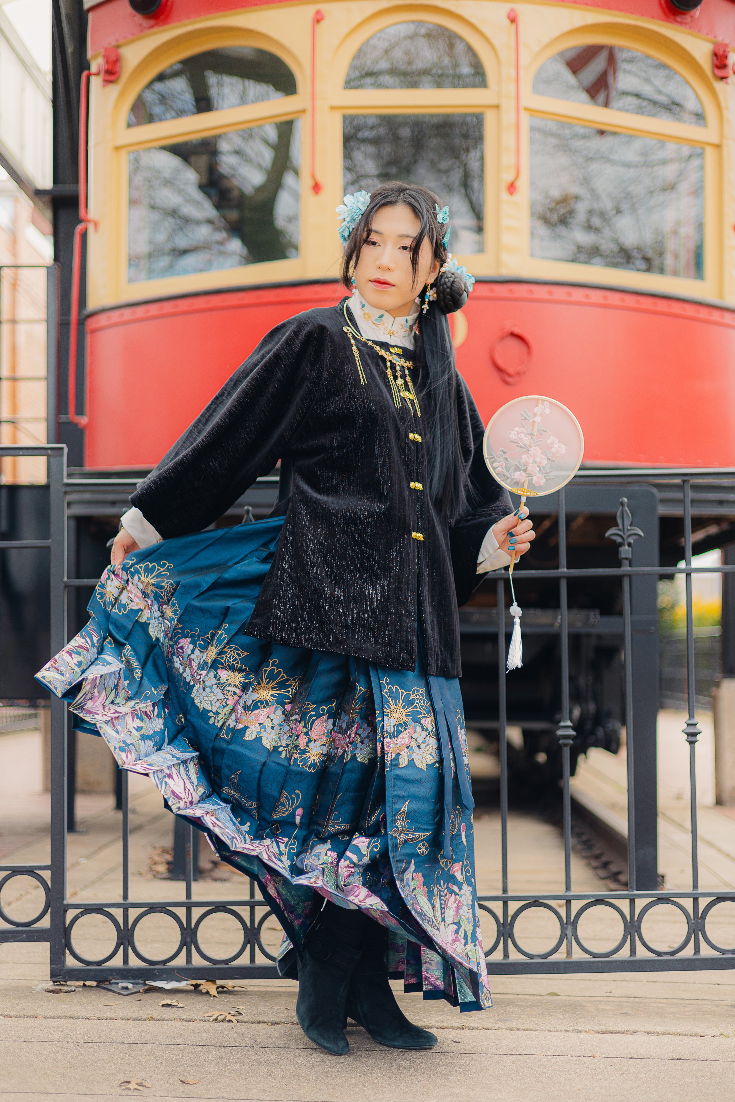 A young woman dressed in traditional Asian attire stands in front of a red and yellow tram, holding a fan with floral design. She wears a dark velvet jacket, a blue floral skirt, and black boots, with her hair styled decorated with flowers.