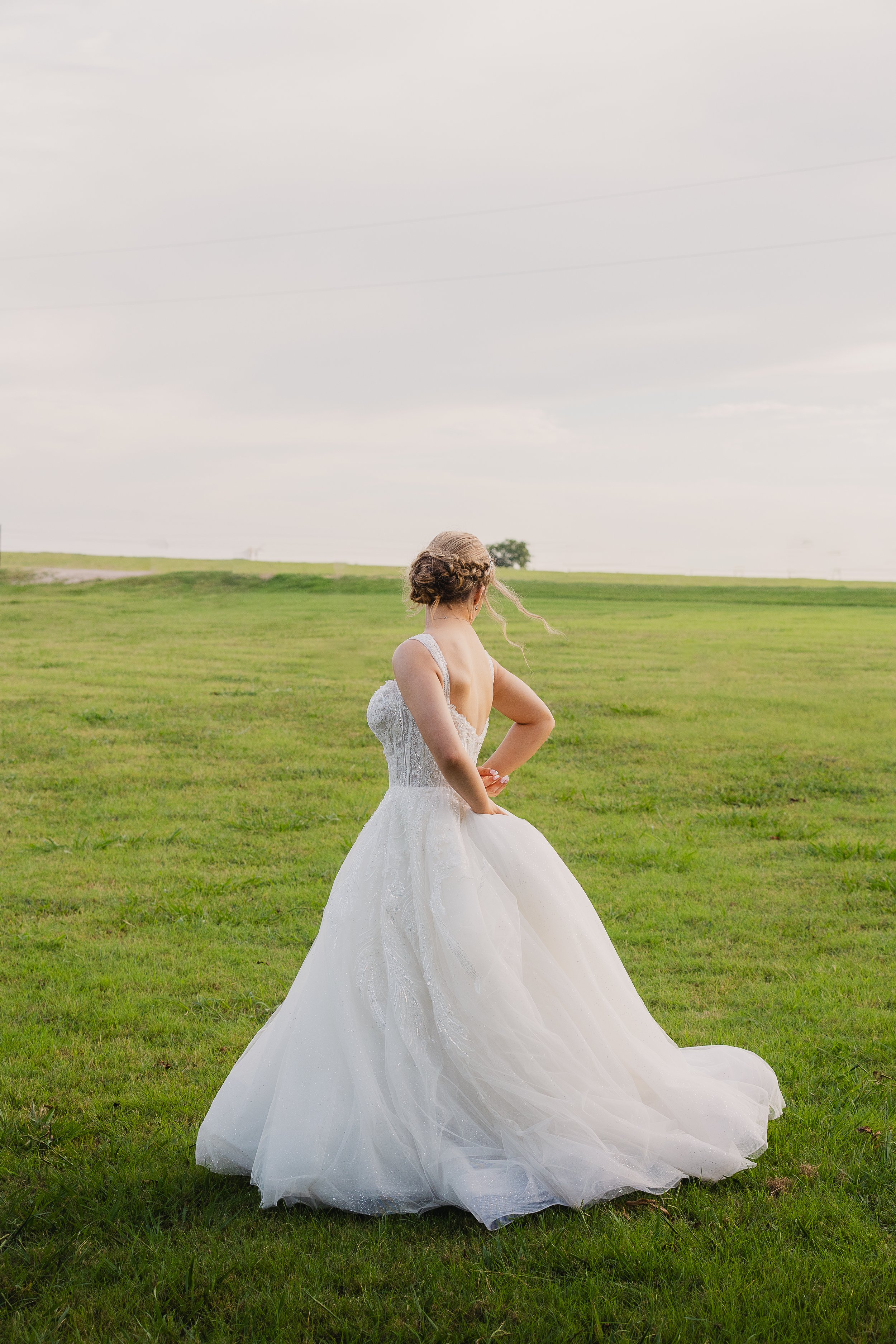 A bride in a white wedding gown standing alone on a grassy field, facing away from the camera, with open sky above.