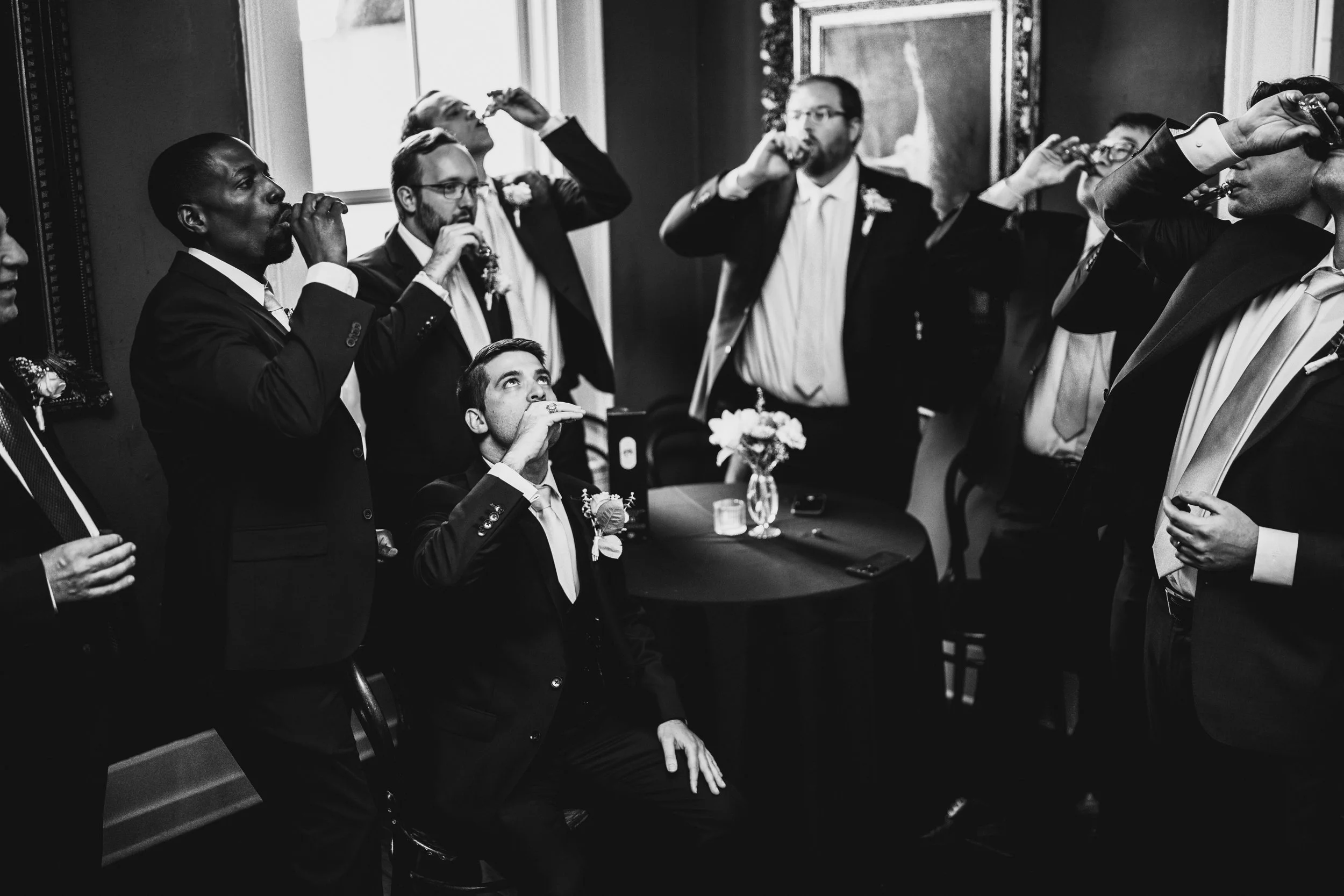 Black and white photo of men in suits taking shots at a gathering, likely a wedding or celebration, with a flower arrangement on a table and framed artwork on the wall in the background.