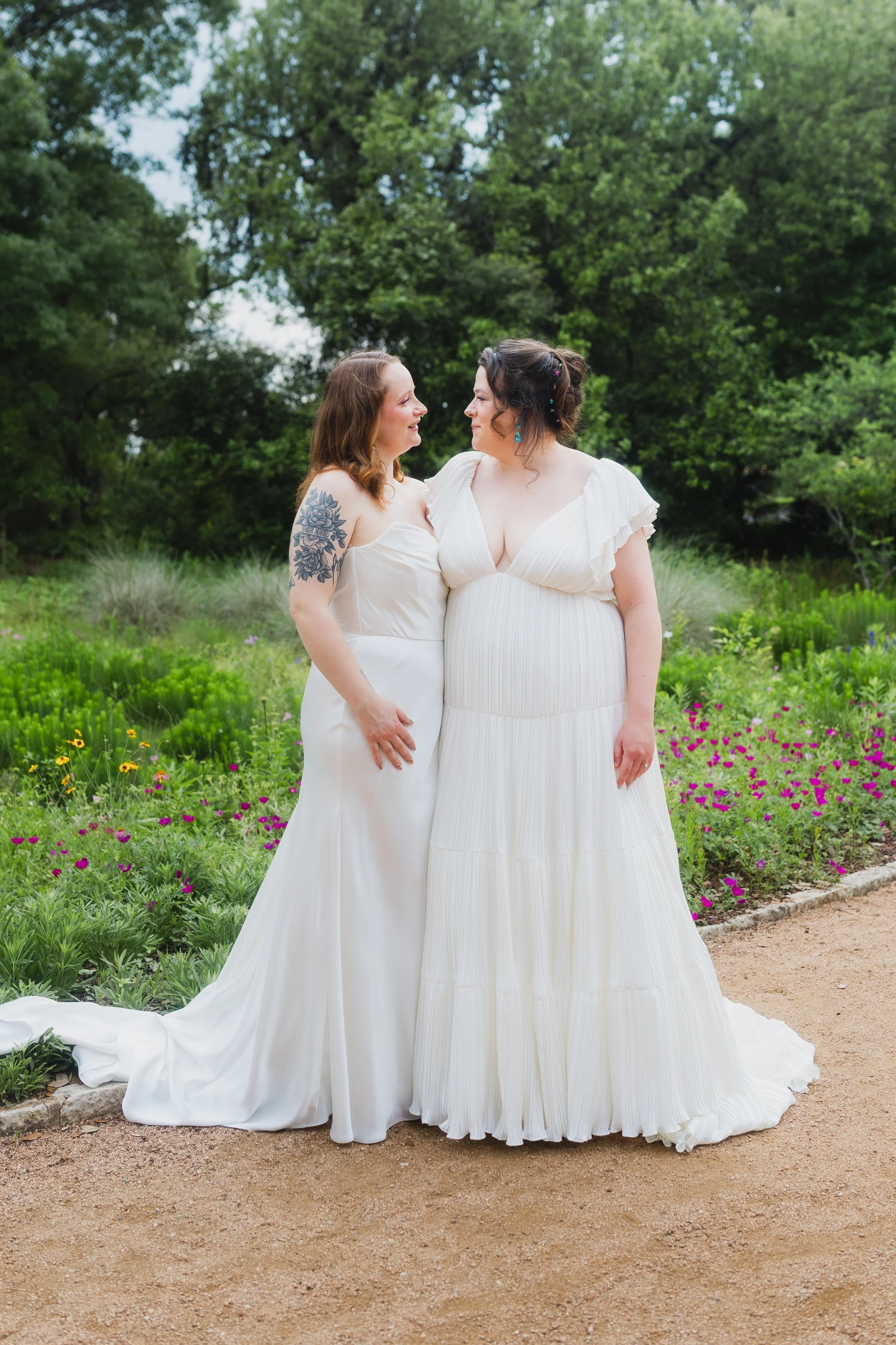 Two women in white wedding dresses standing close and looking at each other in a garden with green trees and purple flowers.