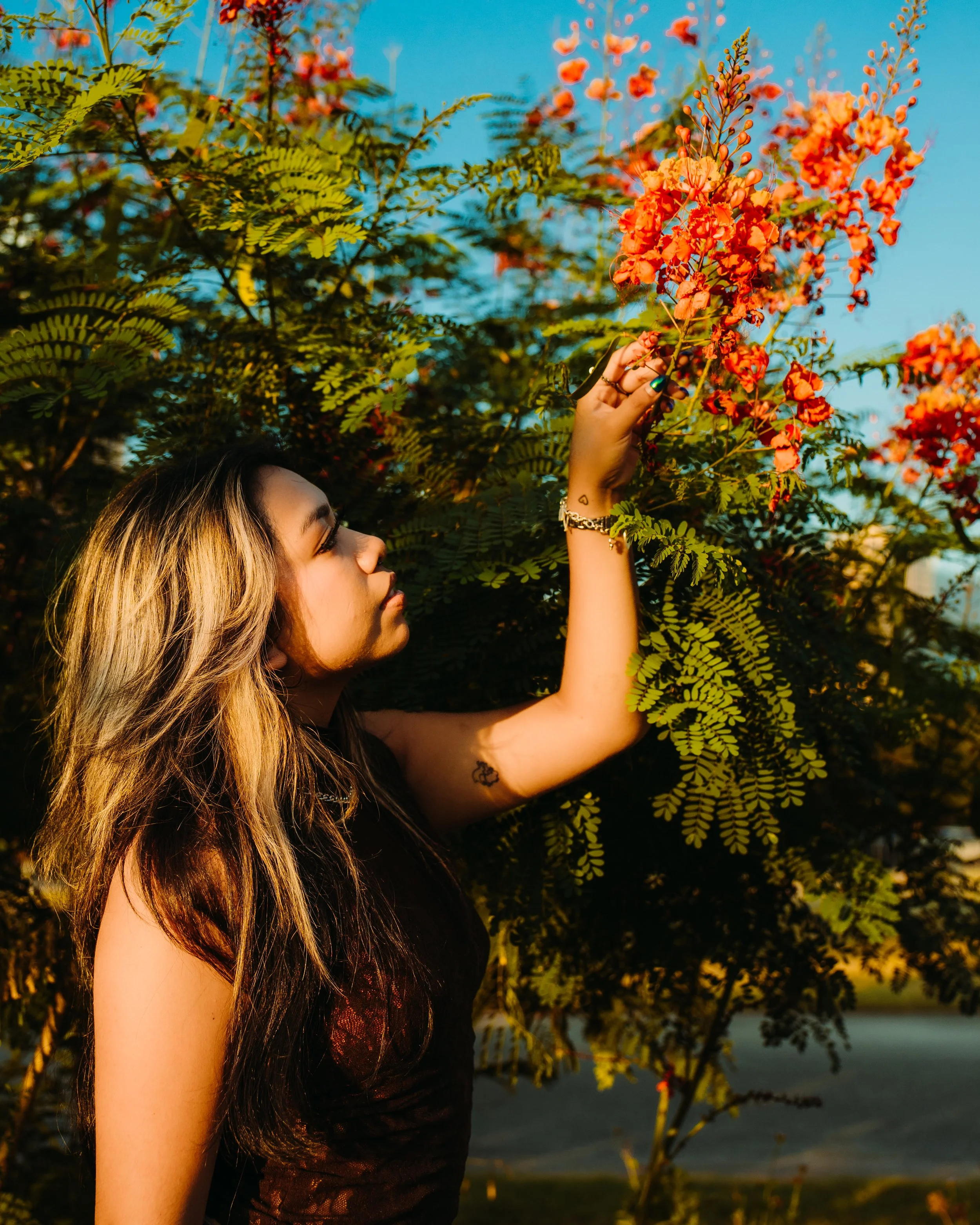 A woman with long, wavy, blonde-brown hair reaching for orange flowers on a bush in sunlight.