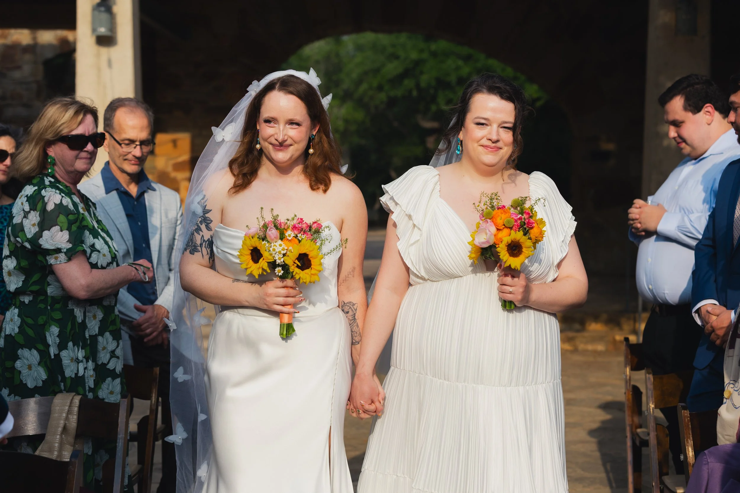 Two brides holding hands and bouquets during a wedding ceremony, with wedding guests in the background.