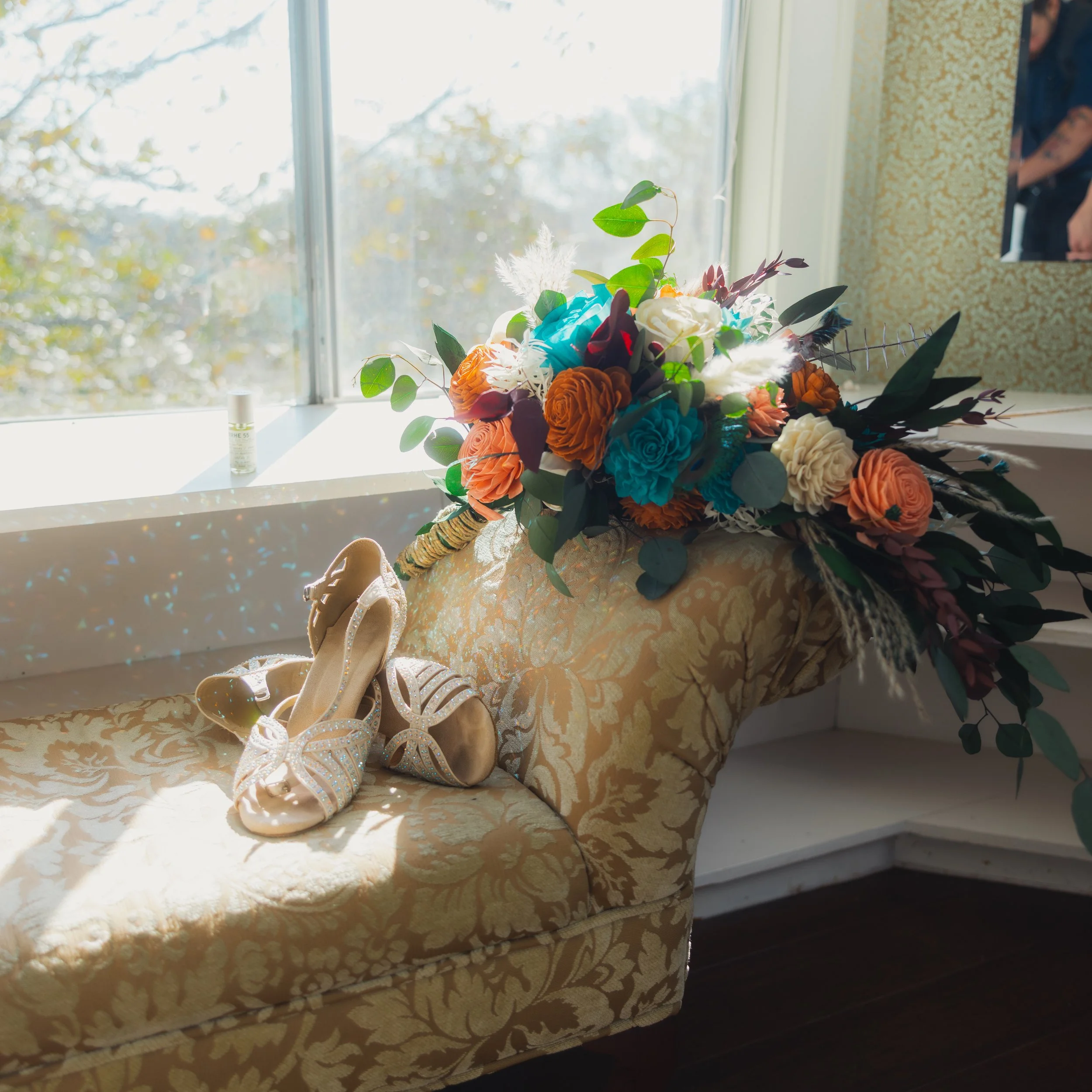 Colorful bouquet of flowers including orange roses, blue roses, white roses, and greenery on a vintage-style, beige patterned sofa. A pair of sparkly high-heeled shoes rests beside the bouquet. Sunlight streams through the window behind, creating a b