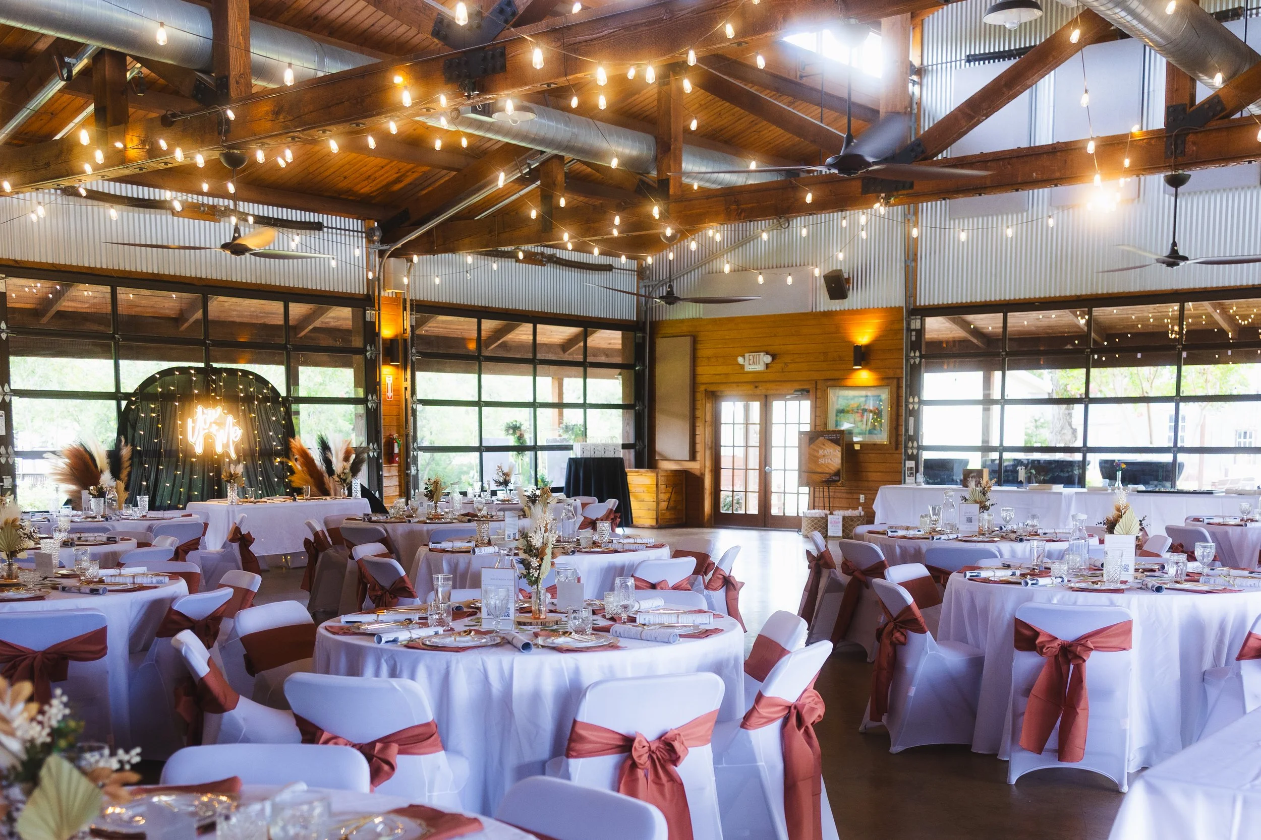 A decorated banquet hall with round tables covered in white tablecloths, adorned with flowers and place settings. Brown sashes are tied around the white chairs. String lights hang from the wooden ceiling beams, and large windows allow natural light t