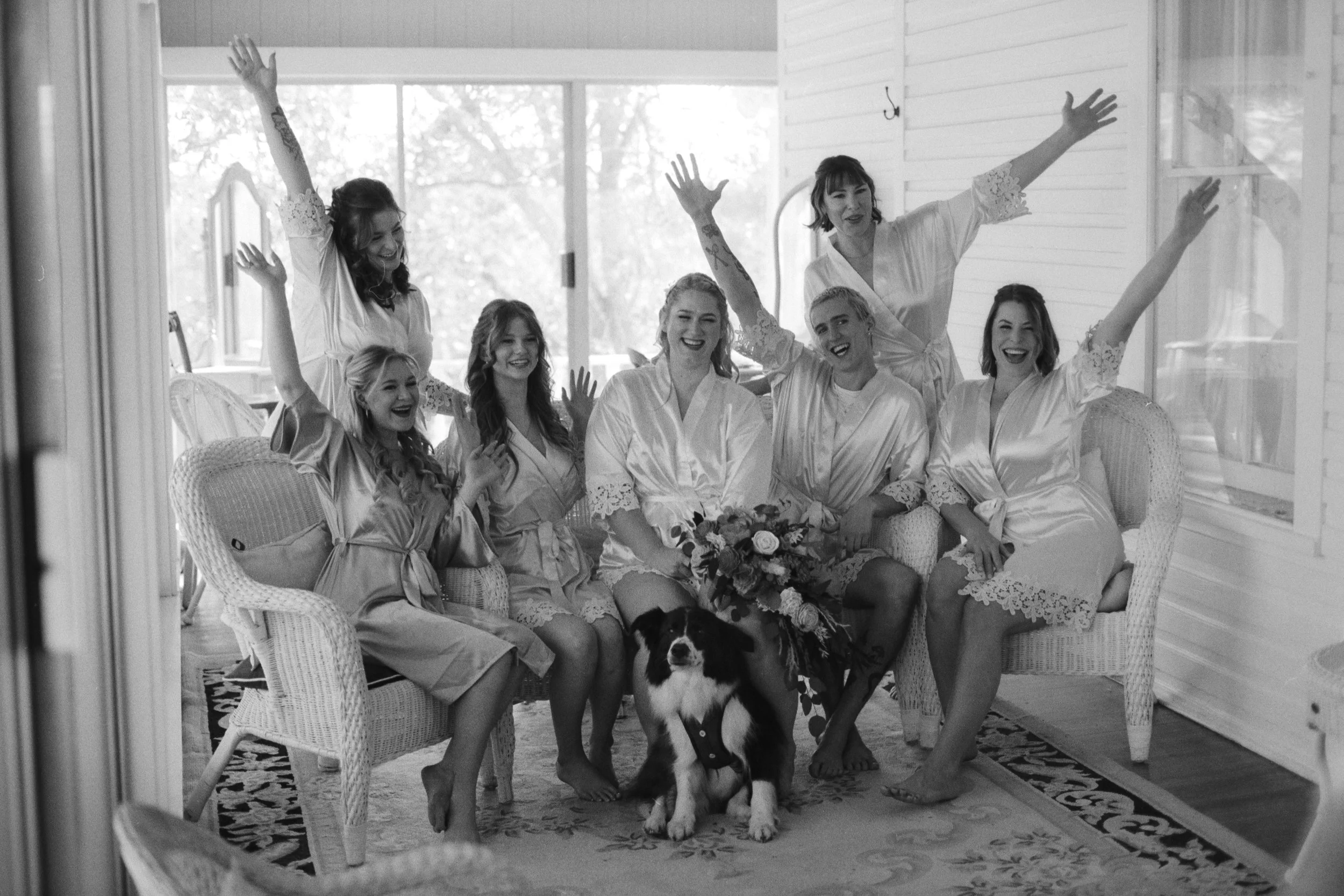 A group of women in satin robes celebrating together, sitting on wicker chairs, with a dog at their feet, inside a sunlit room with large windows.