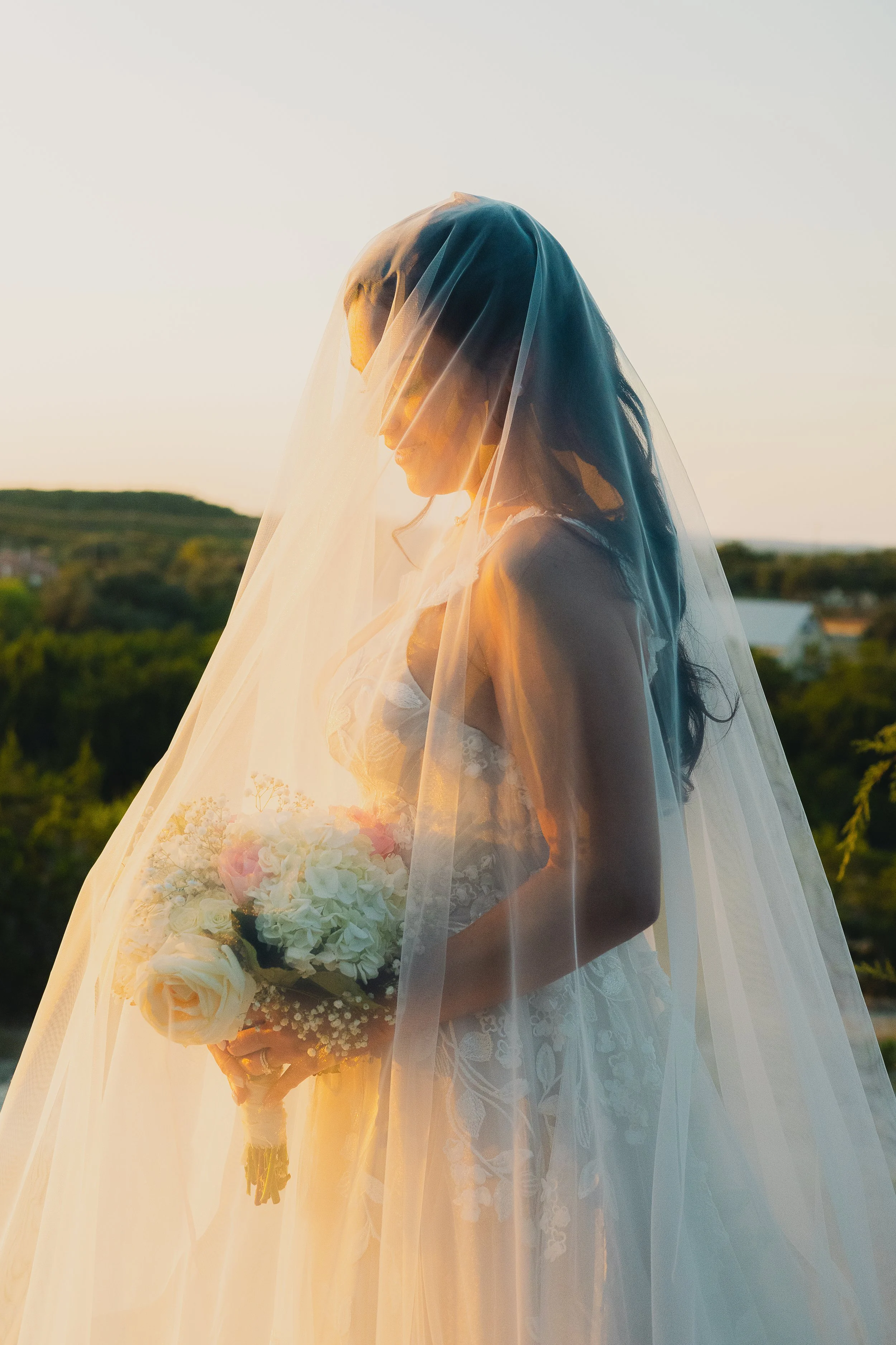 Bride in a wedding dress holding a bouquet, standing outdoors at sunset, with a sheer veil covering her face.