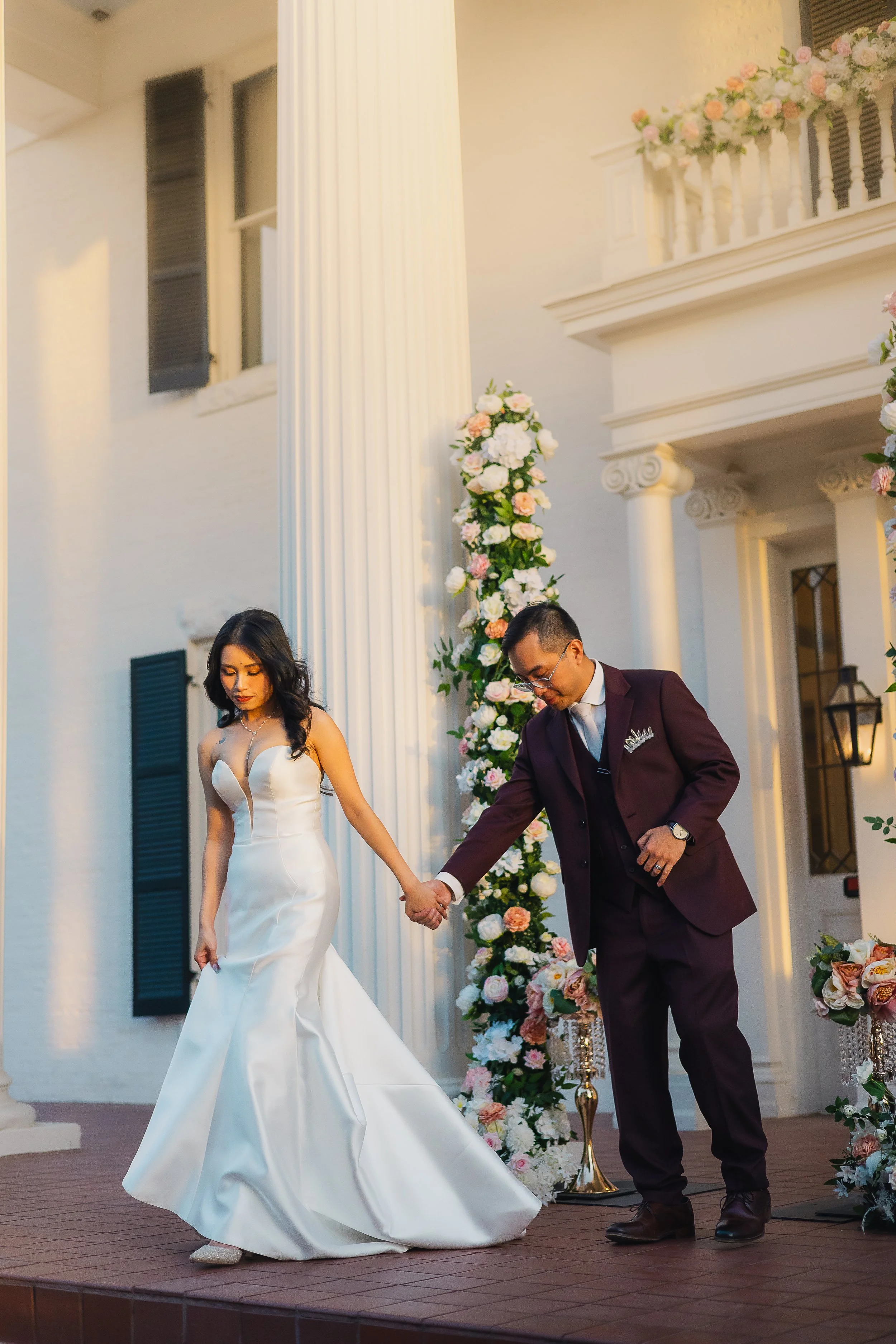 A bride and groom holding hands during a wedding ceremony outside a building with floral decorations.