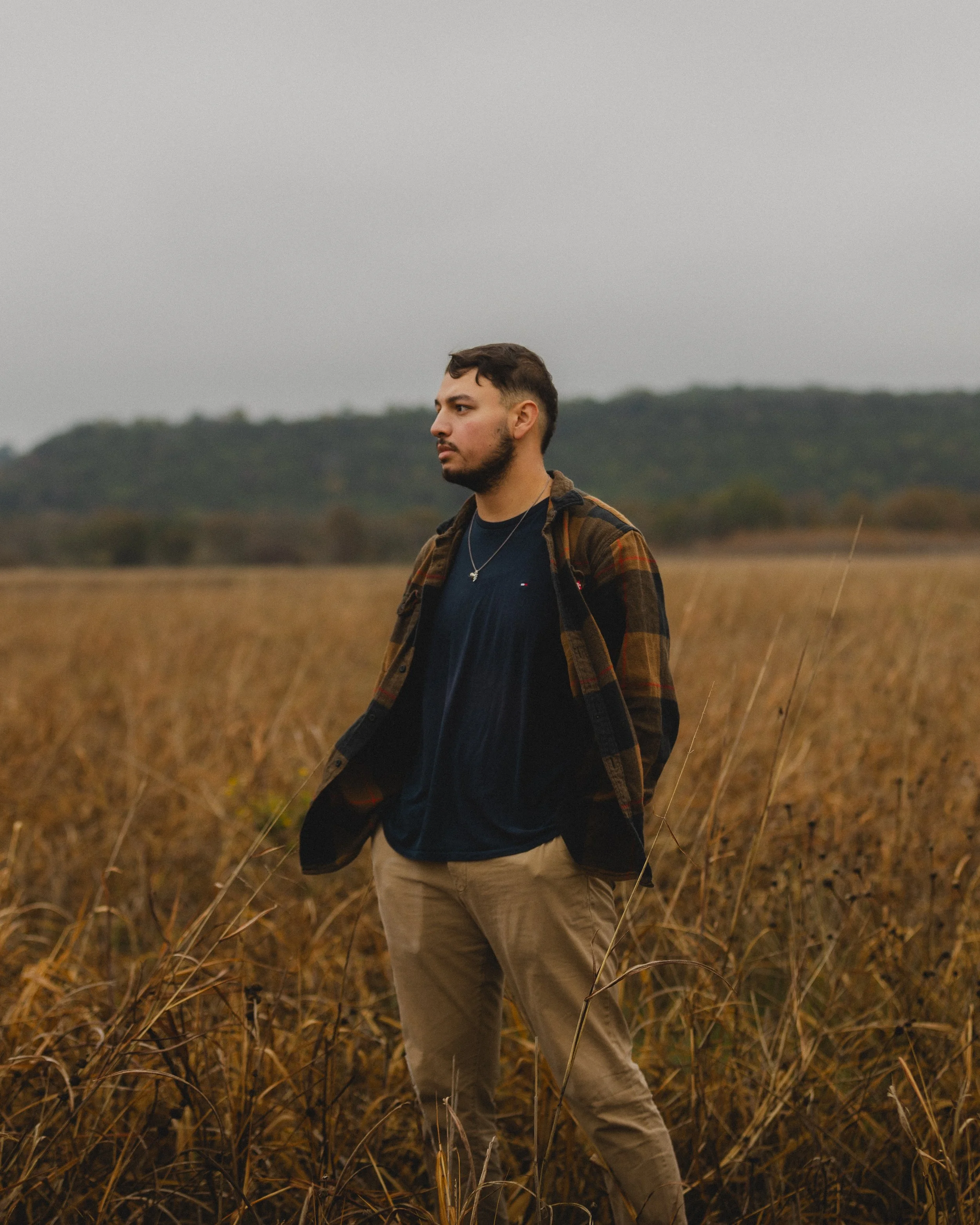 A man standing in a field of tall, dry grass with hills in the background, looking to the side.