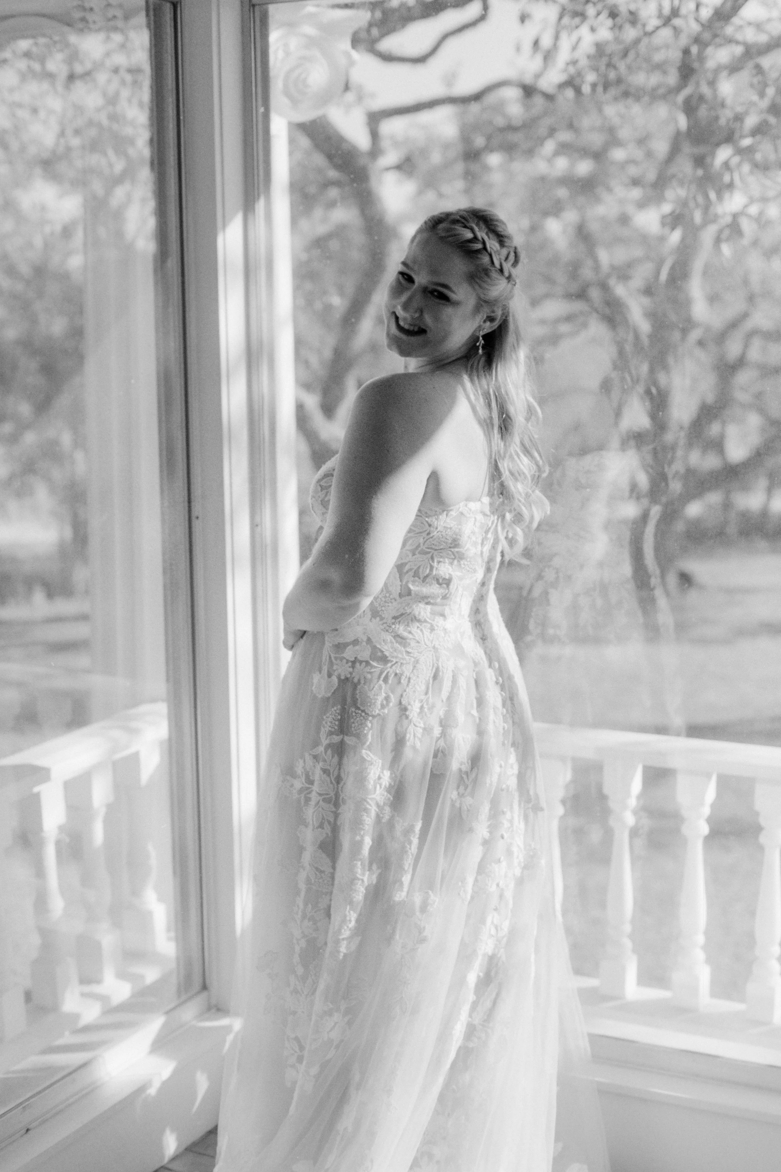 A smiling woman in a strapless, lace wedding dress standing by a large window with trees outside.