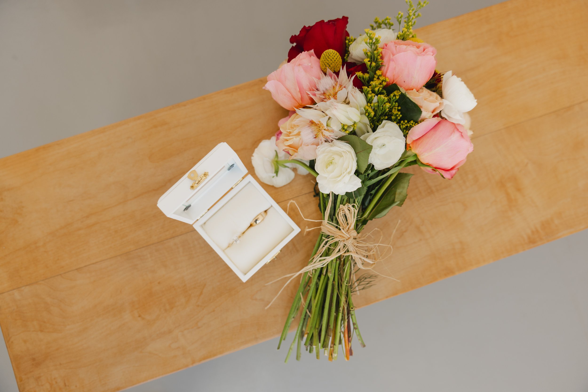 A bouquet of pink, white, red, and cream flowers tied with a raffia ribbon, placed on a wooden surface next to a white ring box with a gold ring inside.