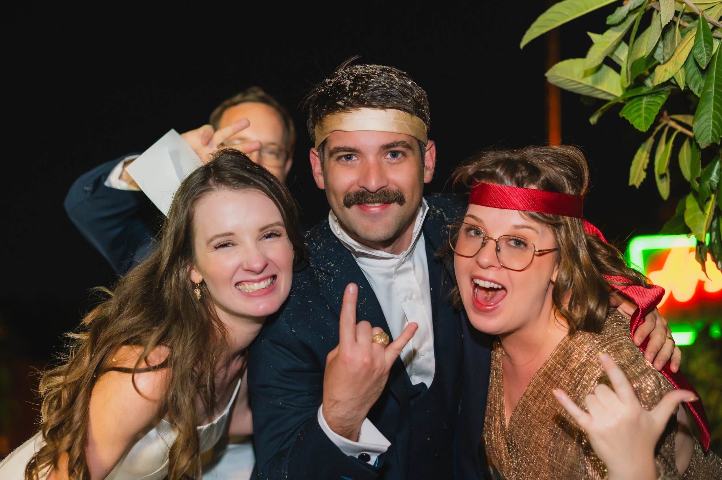 Group of people celebrating at a party, with two women and a man in the foreground making joyful gestures, and a person in the background making a peace sign.