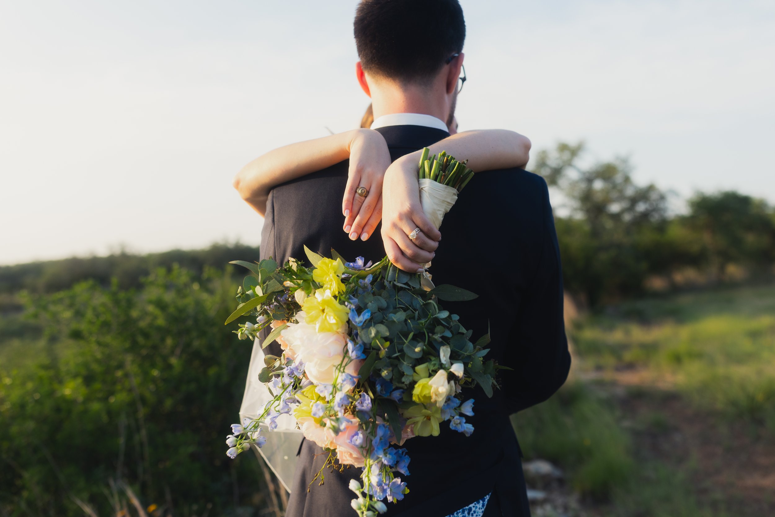 A couple embracing outdoors; the woman holds a bouquet of yellow, blue, and white flowers.