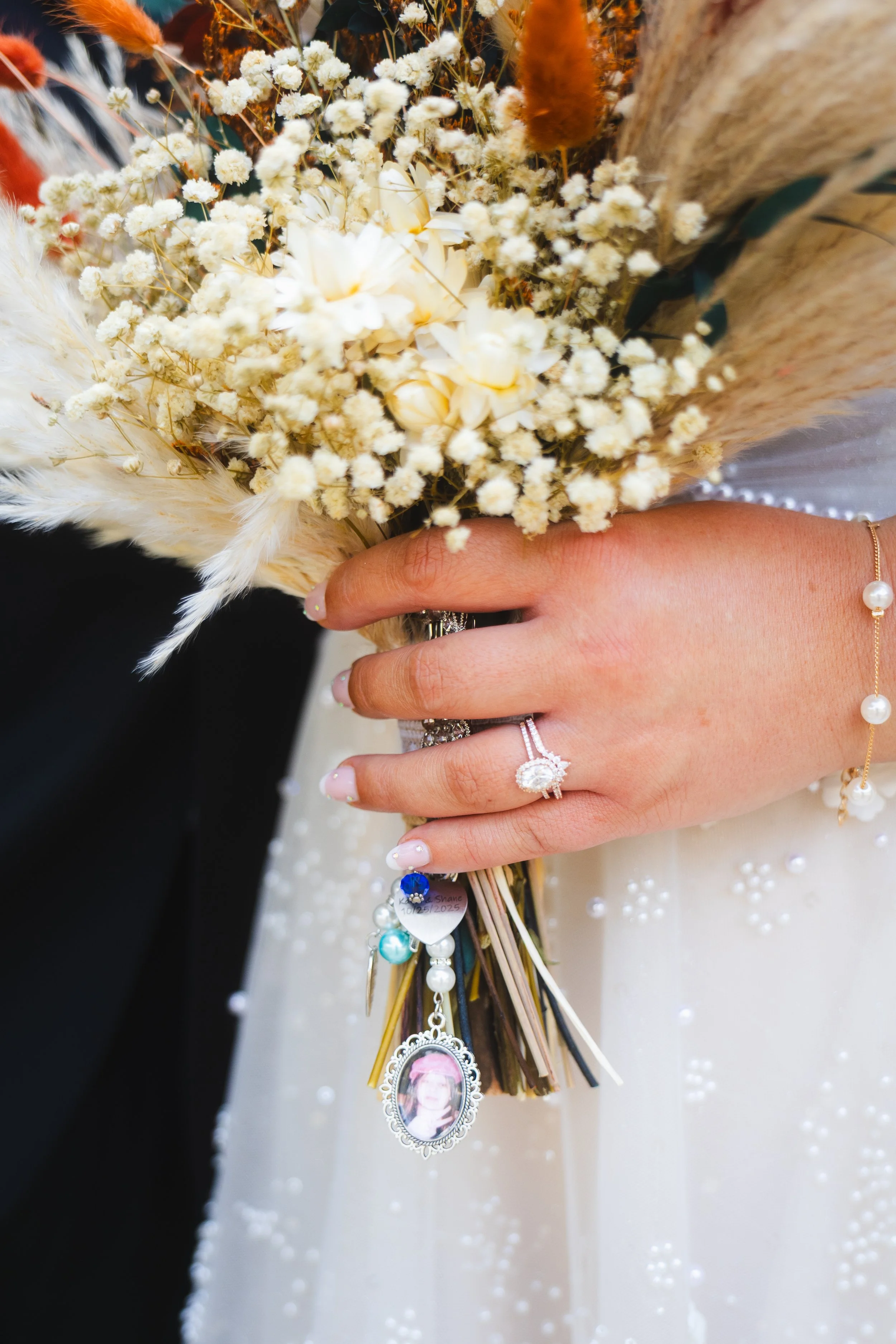 A woman's hand holding a bridal bouquet of white flowers and dried plants, showing rings and jewelry, with a wedding dress decorated with pearls visible in the background.