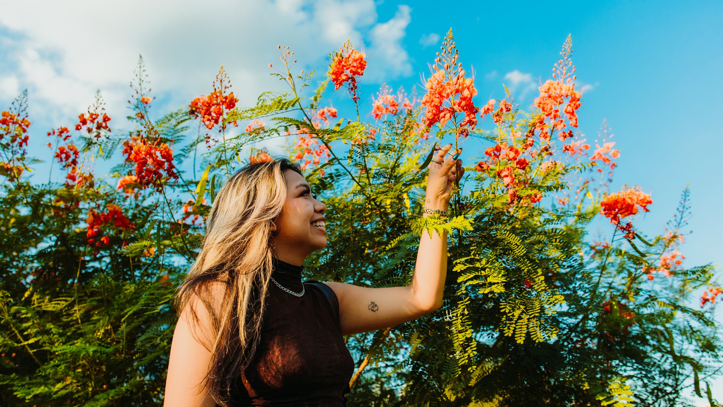 A young woman with long blonde hair smiling as she touches pink flowering branches of a tree outdoors against a blue sky with some clouds.