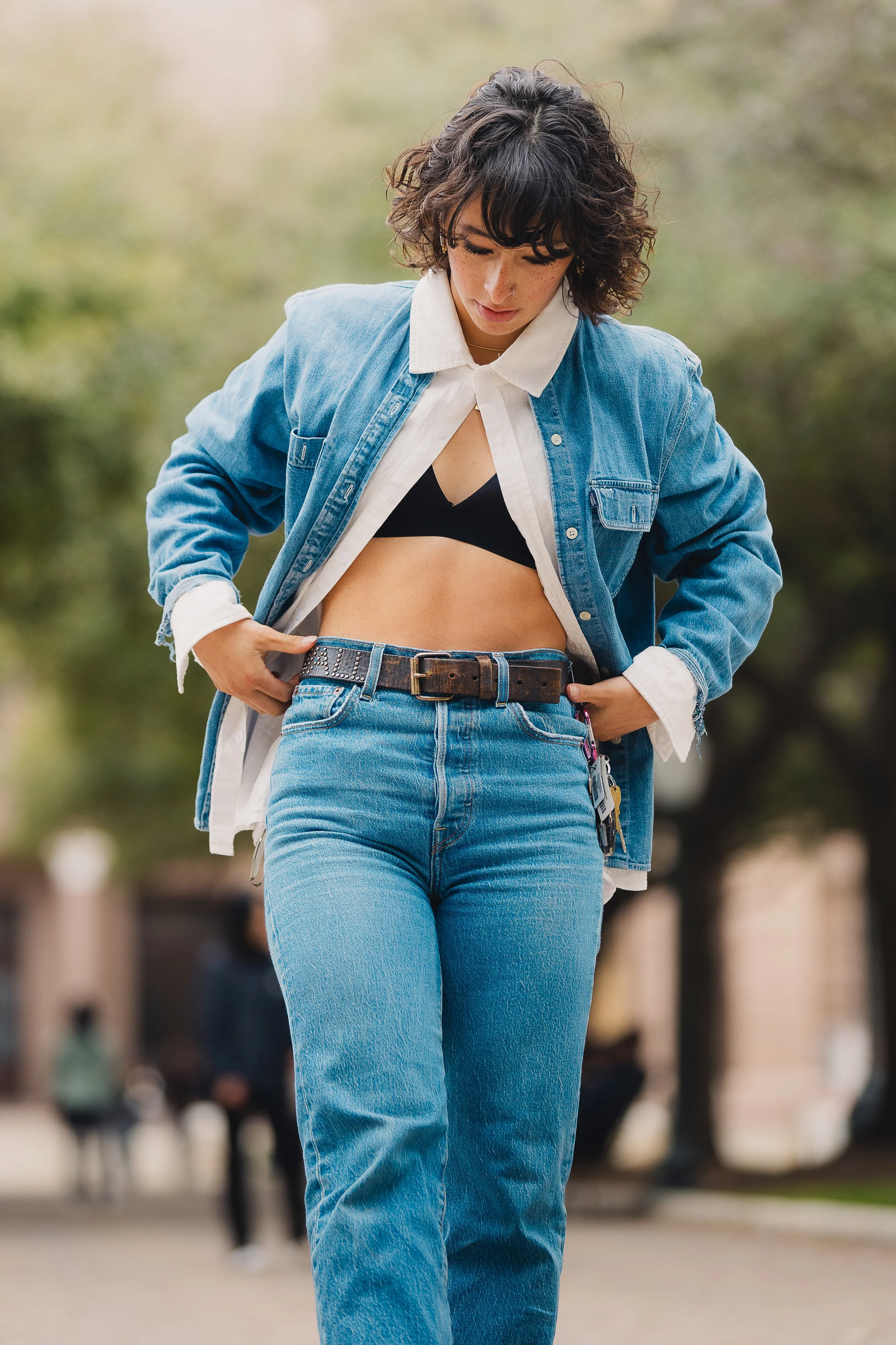 A woman wearing a denim jacket and jeans, with a black crop top, adjusting her belt, outdoors with blurred trees and people in the background.