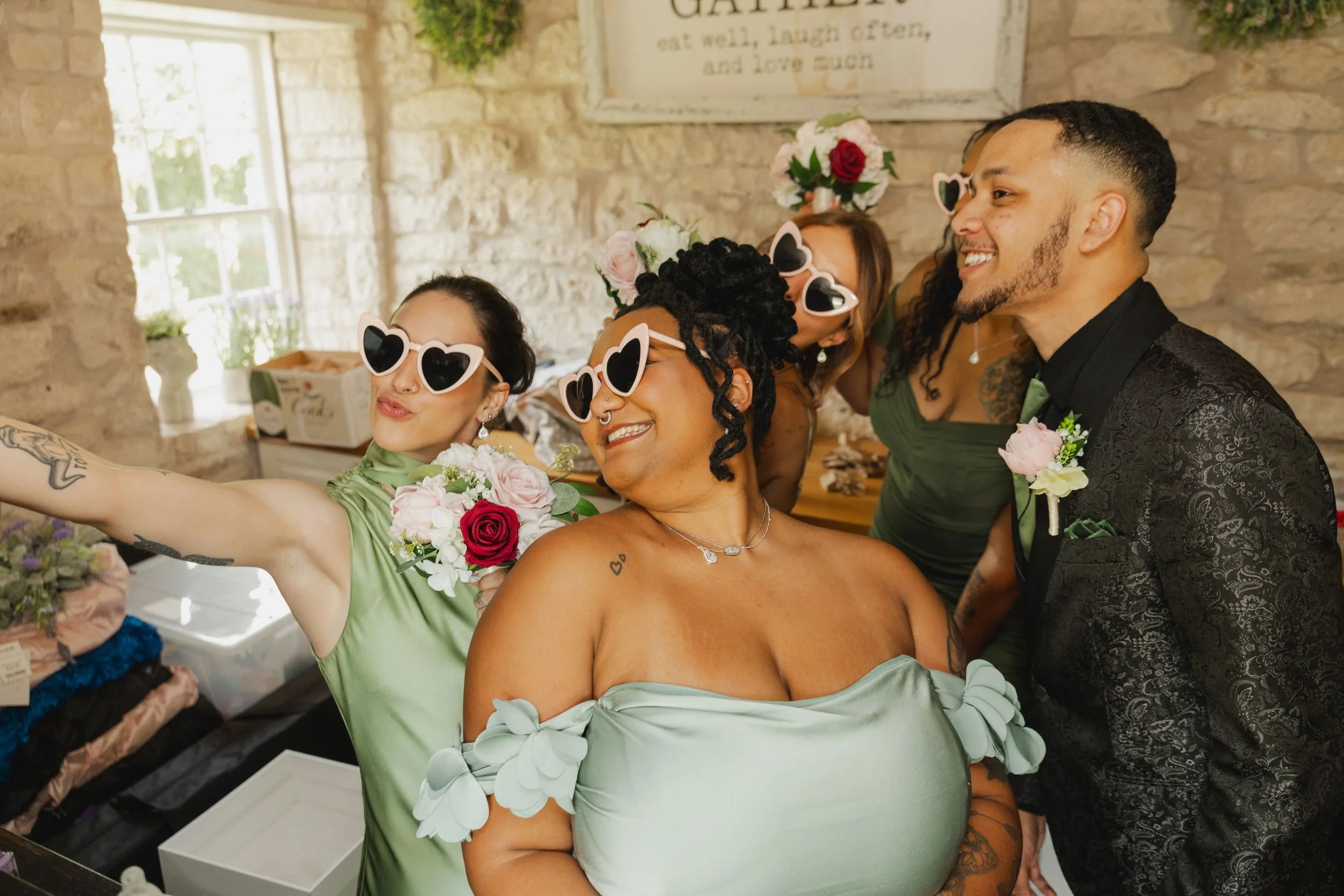 A group of people at a wedding, taking a selfie. They are smiling, wearing heart-shaped sunglasses, with some holding bouquets of flowers. The background shows a rustic stone wall and window, with a sign above reading 'Gather, eat well, laugh often, 