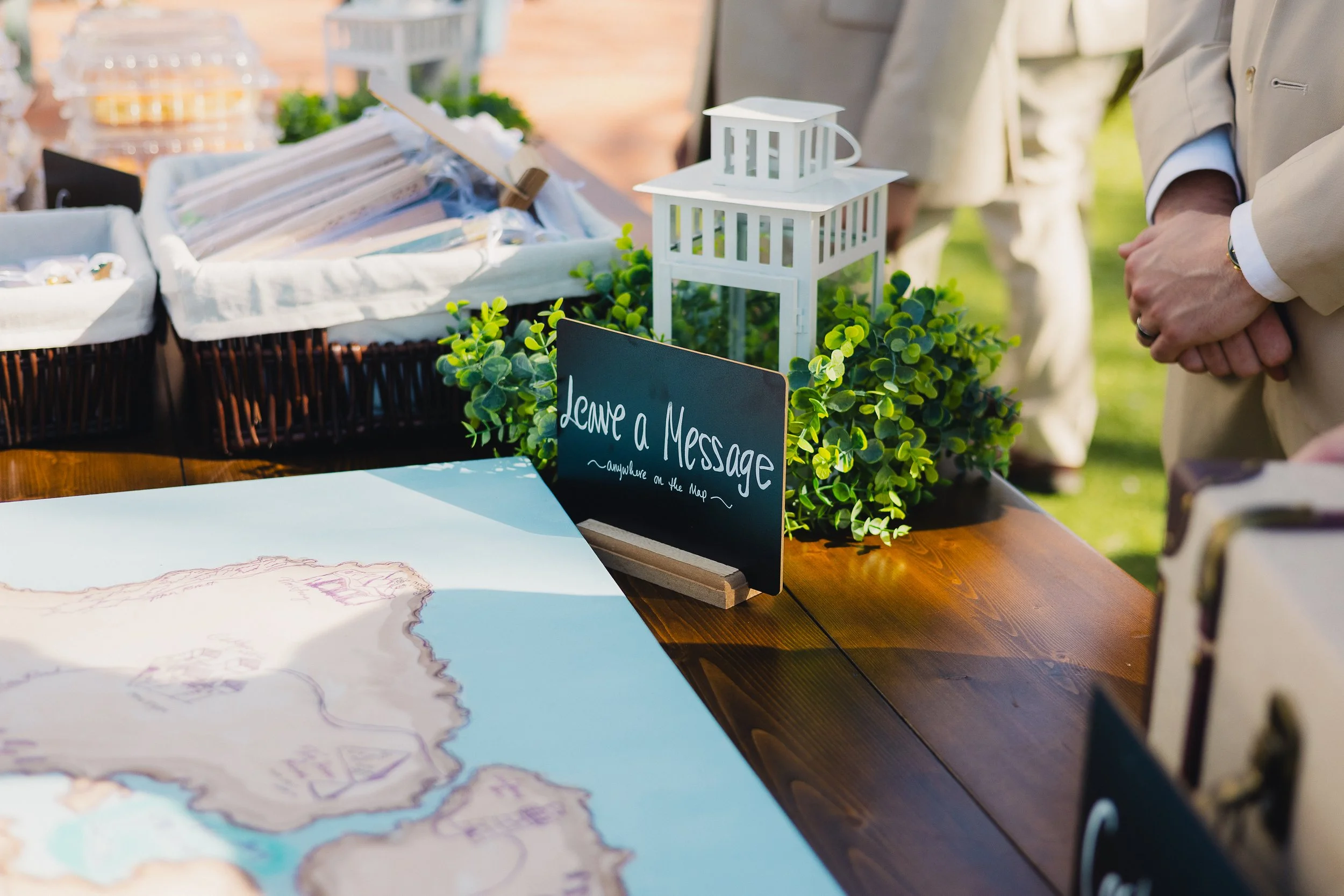 A table at an outdoor event with a small blackboard sign that reads 'Leave a Message' next to green plants, lantern decor, and a map.