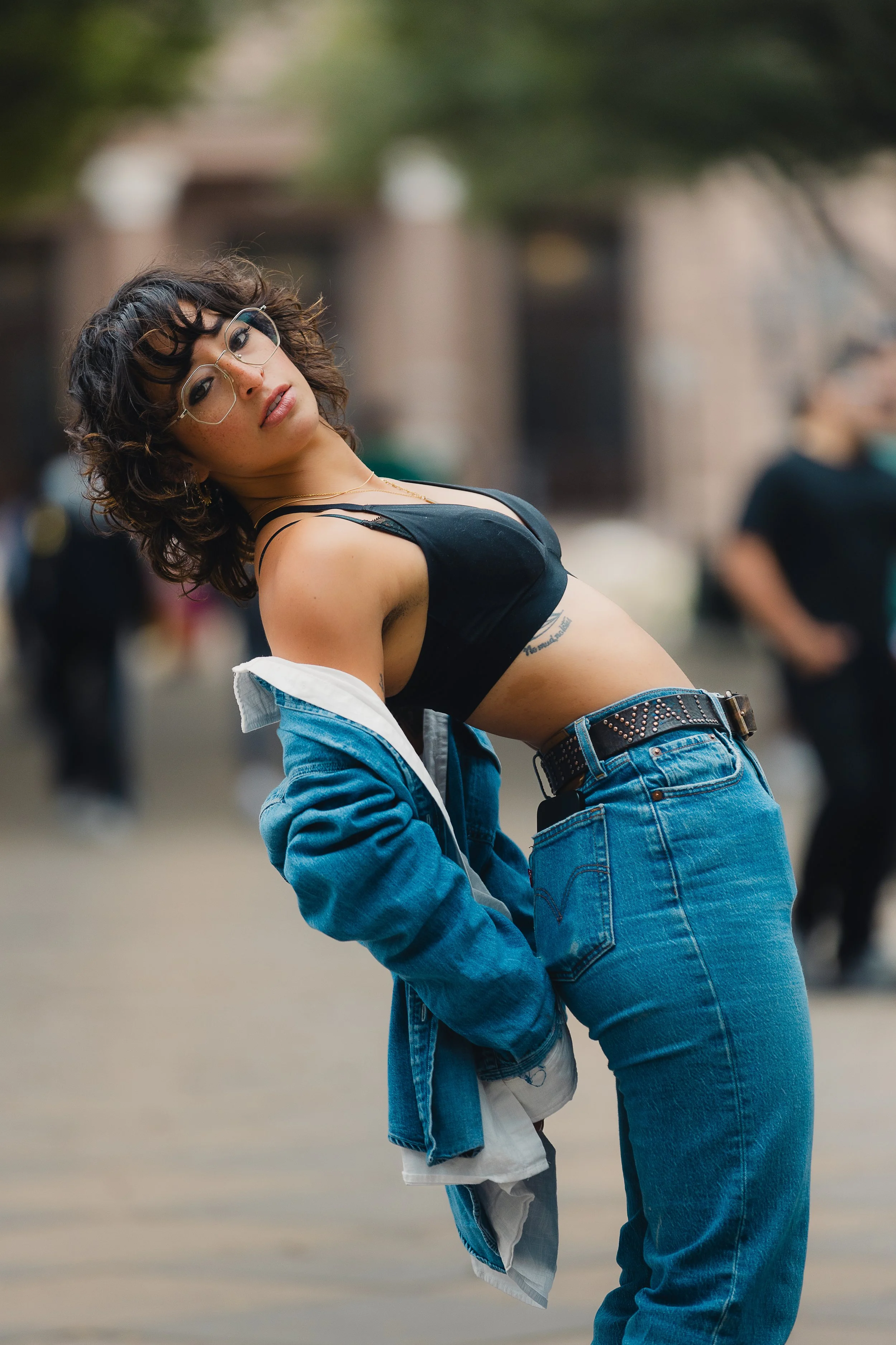 A woman with dark curly hair, wearing glasses, a black crop top, blue jeans, and an open denim jacket, posing outdoors with a blurred background.