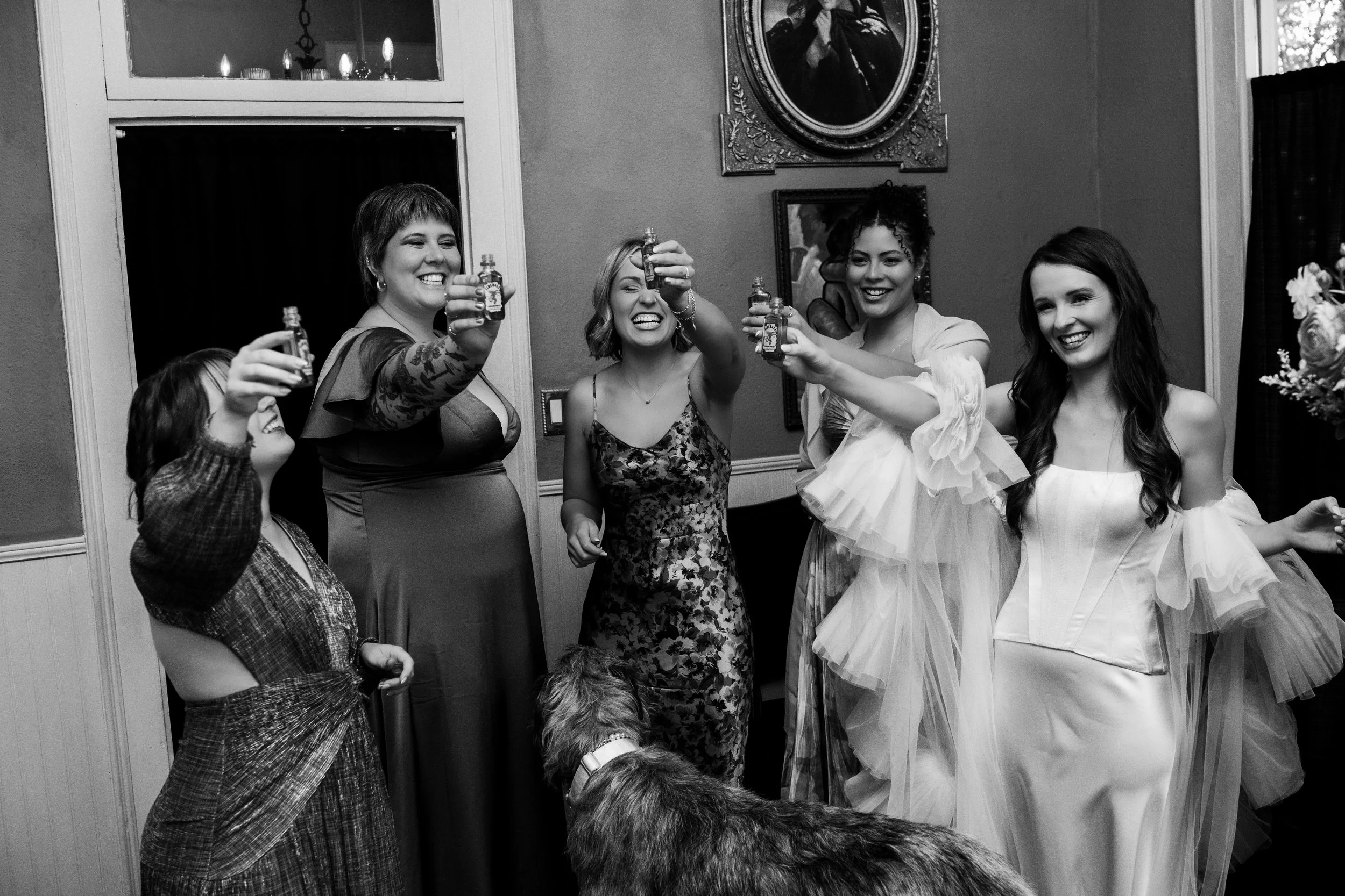 Group of women at a celebration raising small bottles together, some smiling and laughing, in a decorated indoor setting.