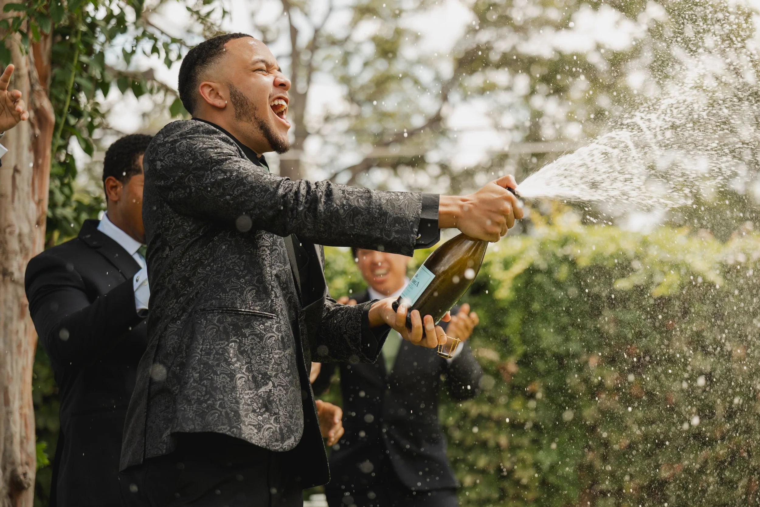 A group of men celebrating outdoors, with one man opening a champagne bottle and spraying it in the air. They are dressed in suits and appear to be happy and excited.