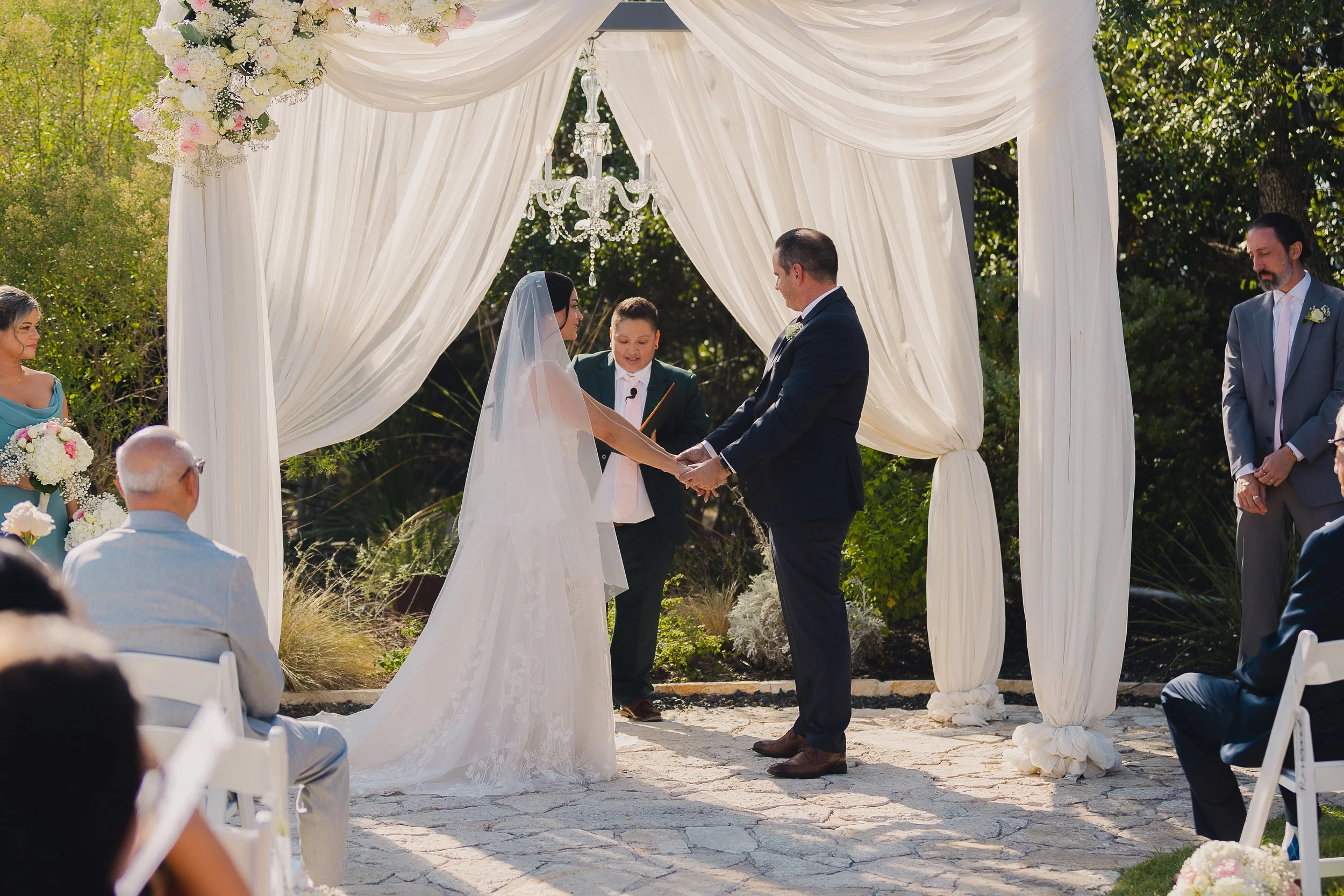 A wedding ceremony outdoors with a bride and groom holding hands under a white draped arch, surrounded by guests and floral arrangements, during daytime.