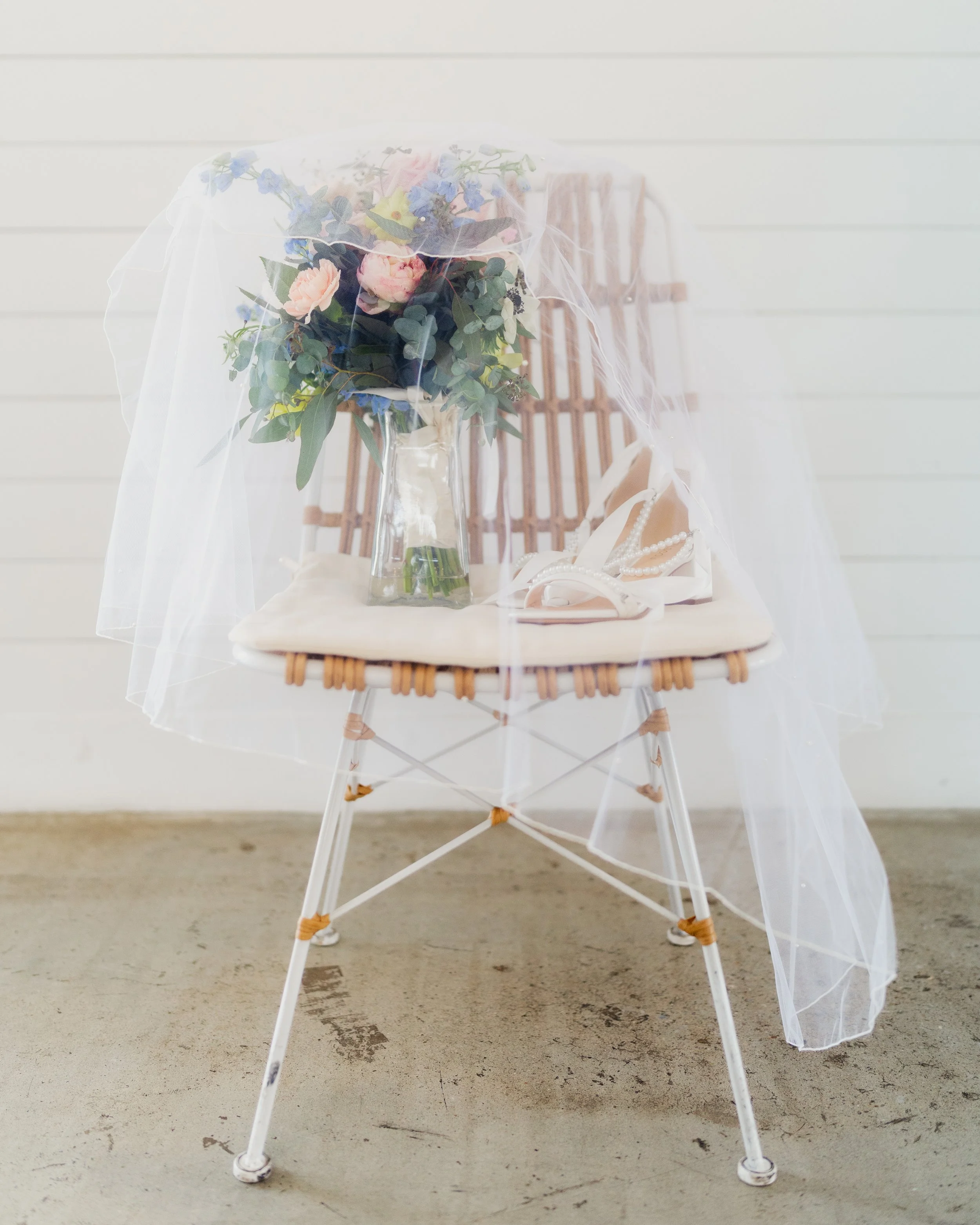 A chair with a bouquet of flowers, a pair of white high-heeled shoes, and a veil draped over it, set against a white wooden wall.