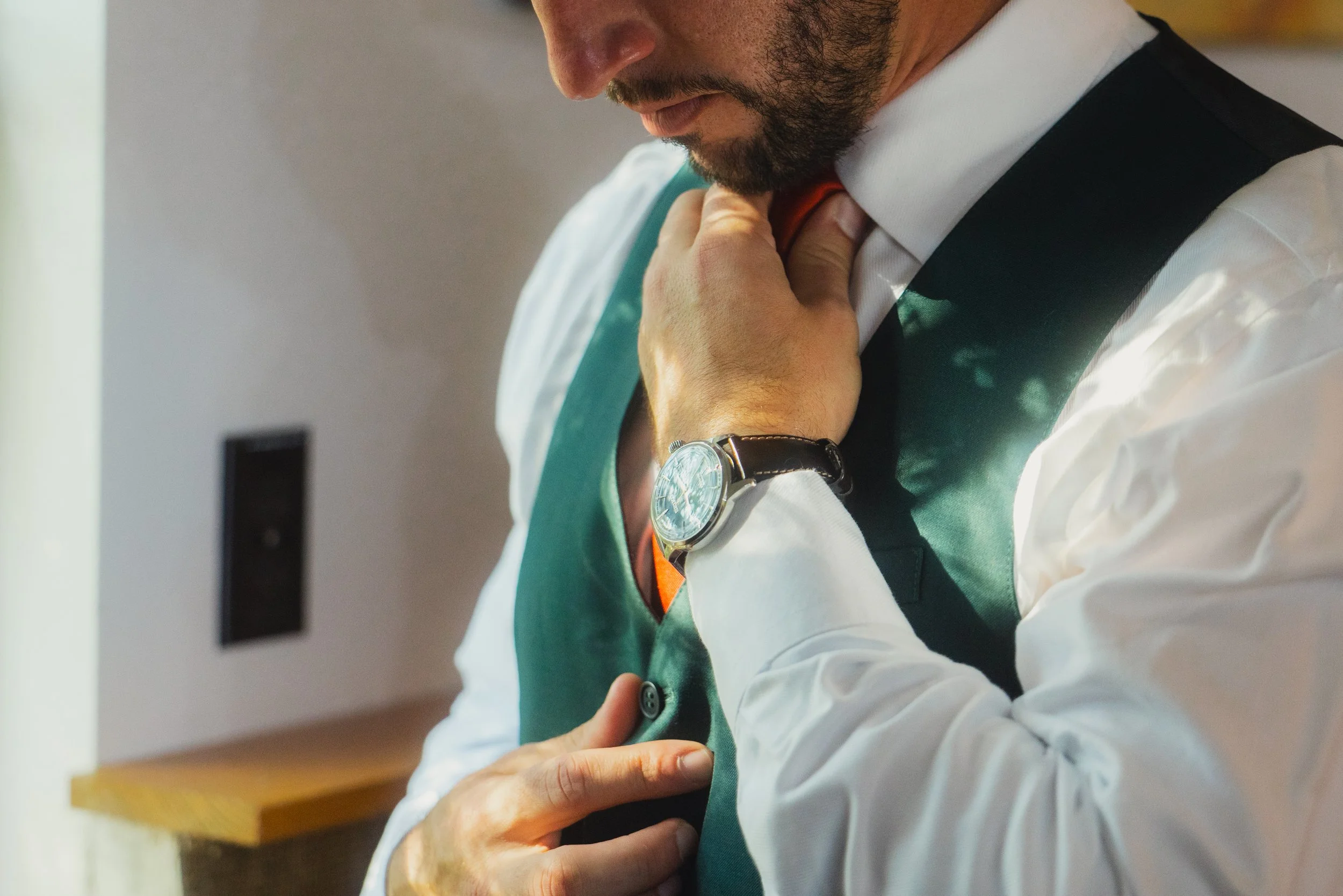 Close-up of a man adjusting his tie, wearing a white dress shirt, green vest, and wristwatch.