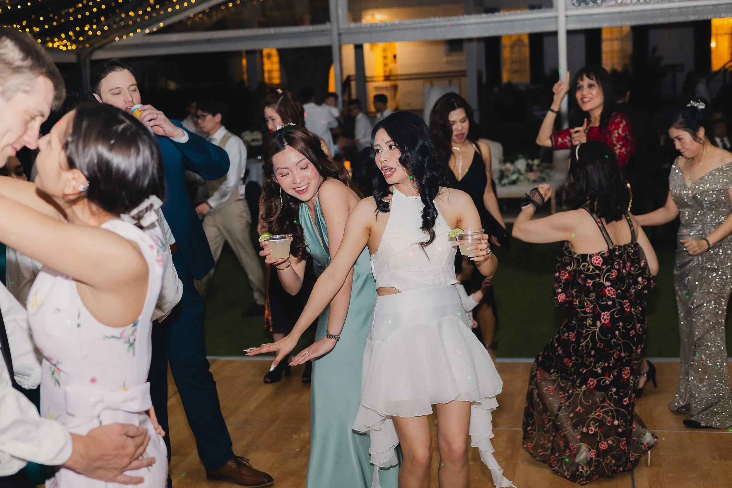 People dancing and enjoying a celebration or party in elegant attire, with some holding drinks, in an outdoor or semi-outdoor setting with string lights overhead.