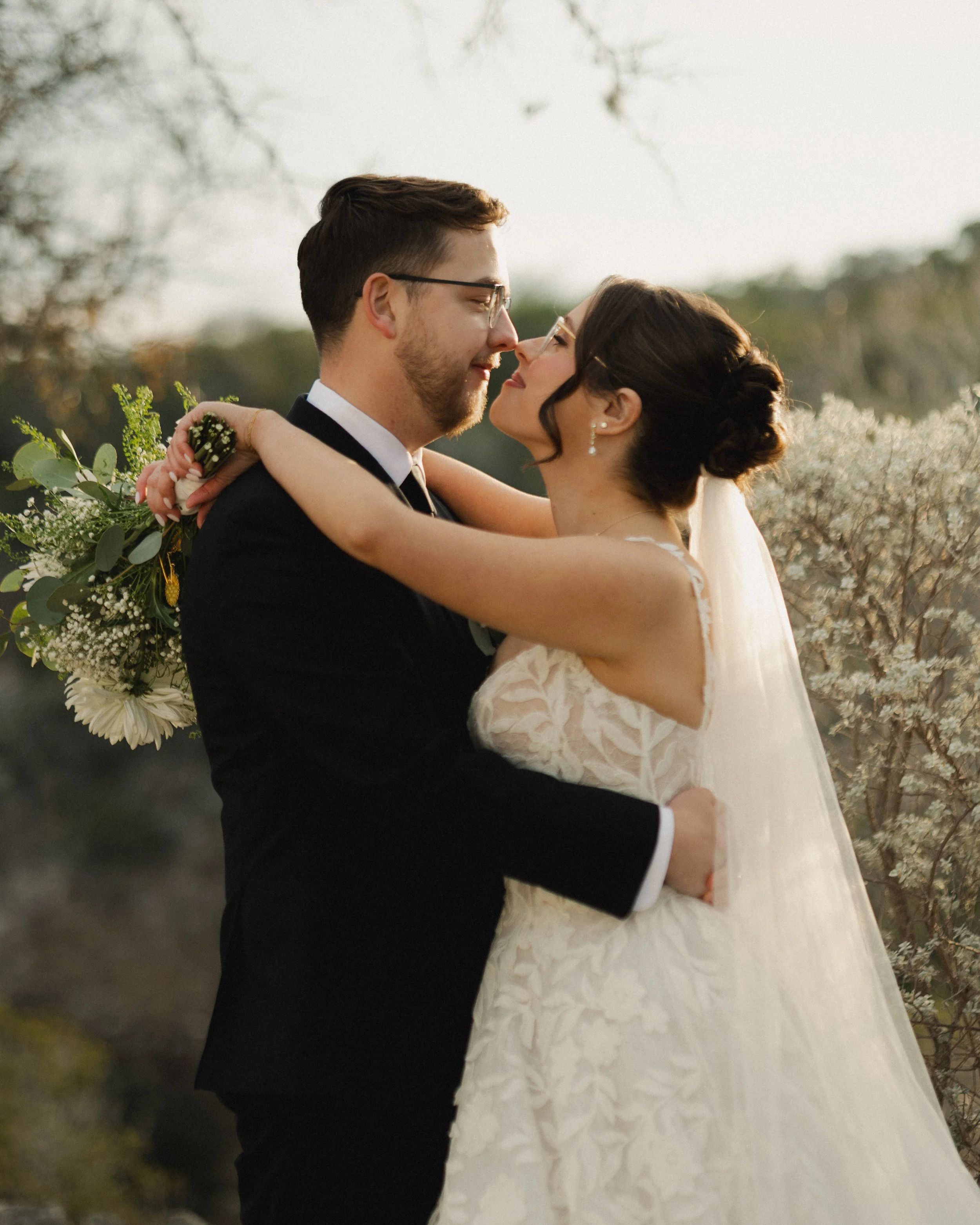 A bride and groom embrace outdoors during sunset, with the groom holding a bouquet of white flowers and greenery. They are smiling and looking into each other's eyes amid flowering trees and soft natural light.