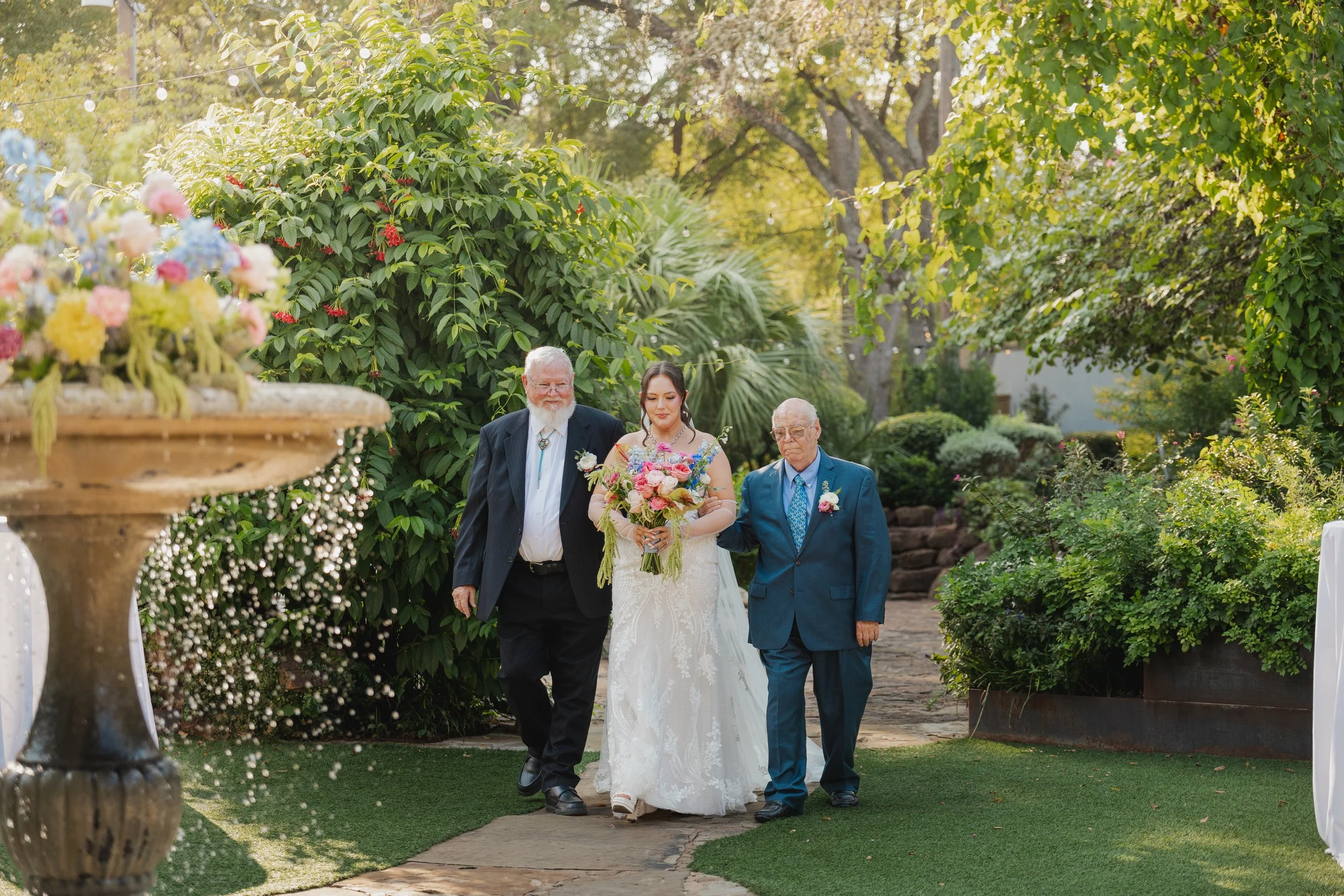 A bride in a white wedding gown holding a colorful bouquet, walking outdoors with two elderly men on either side, surrounded by lush green trees and plants in a garden setting.