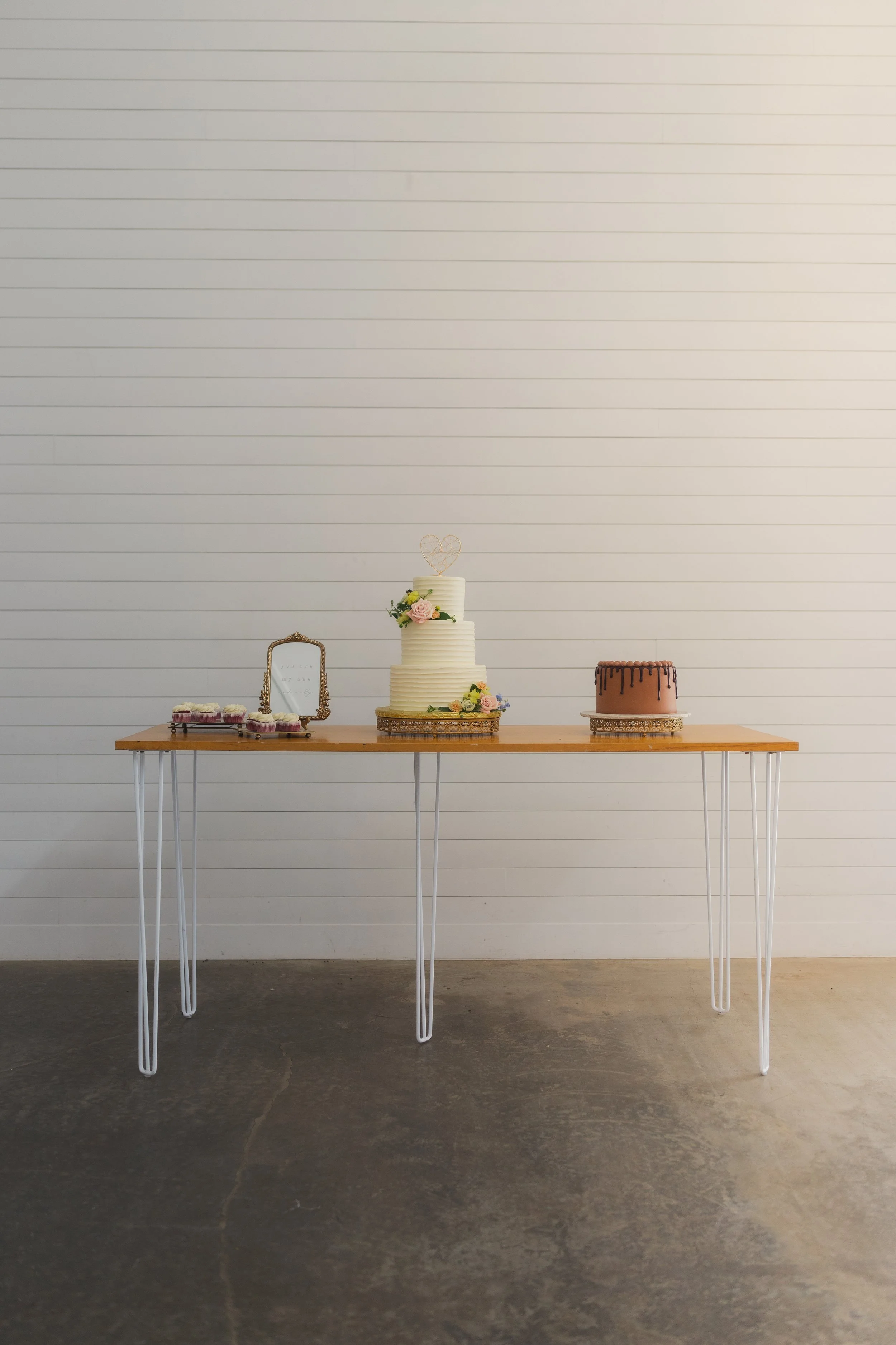 A wooden table with white metal hairpin legs against a white paneled wall, holding a wedding cake, a smaller cake, cupcakes, and a small mirror.