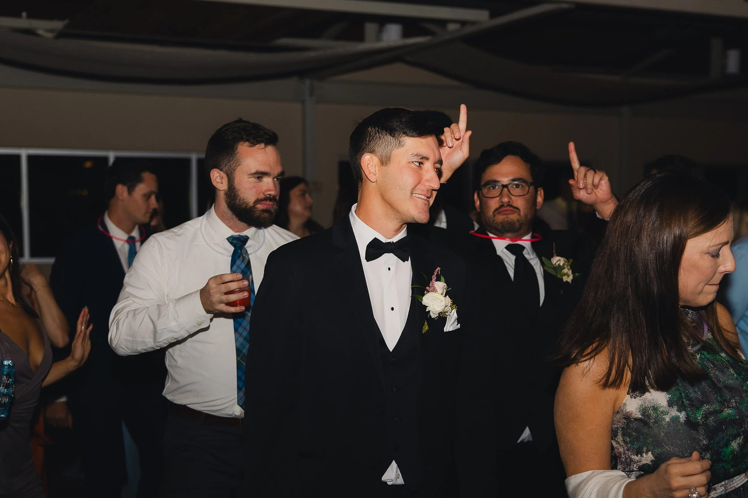 Group of people at a wedding reception, with a man in a tuxedo smiling and raising one finger, others around him holding drinks and dancing.