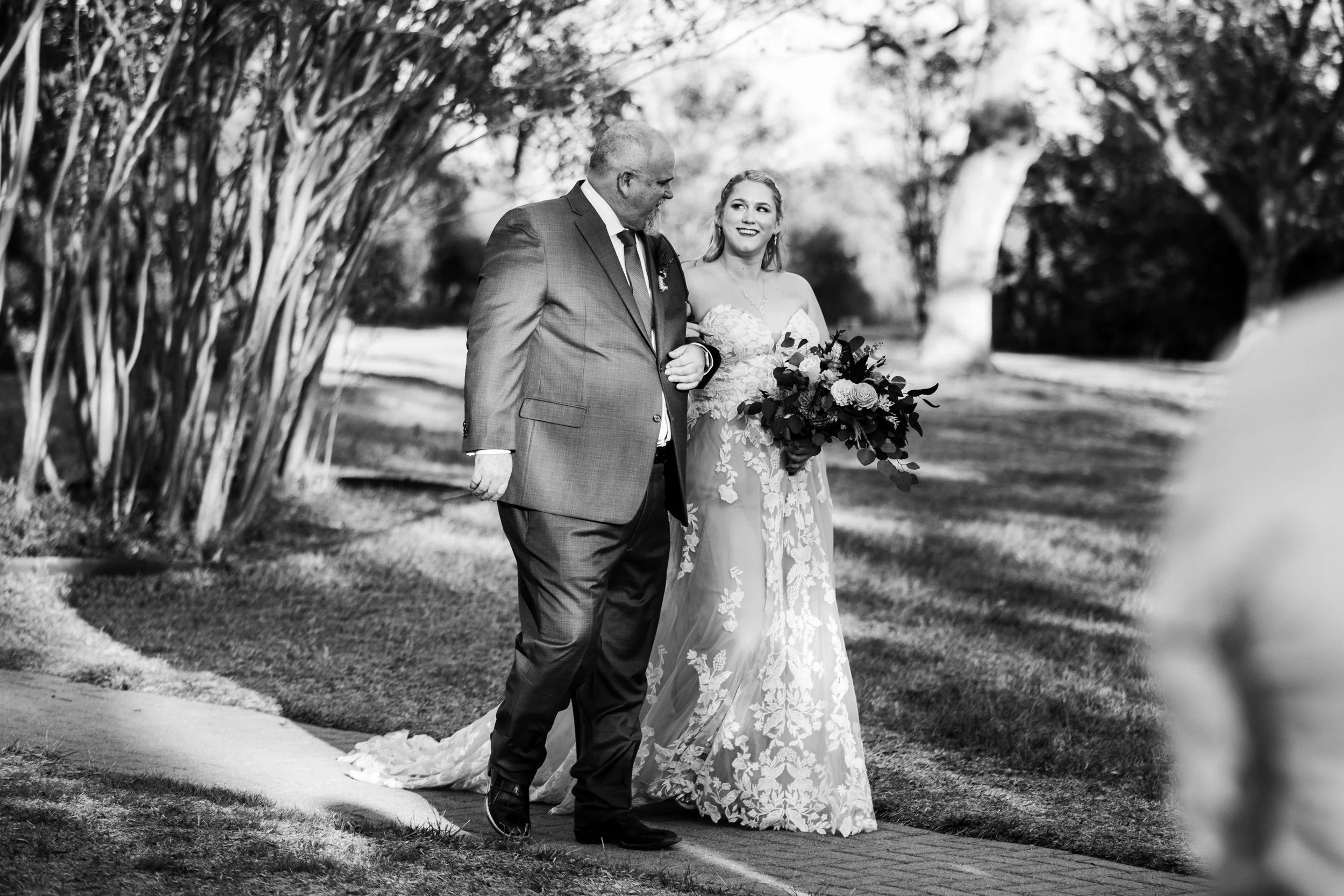 A bride with a bouquet of flowers walking with an older man outdoors, trees in the background, black and white photograph.