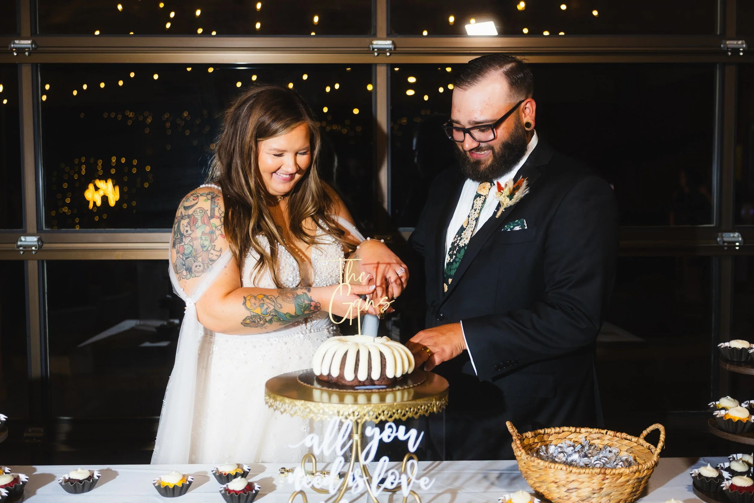 A newlywed couple with tattoos celebrating at their wedding reception, cutting their wedding cake together amid cupcakes and a 'all you need is love' sign.