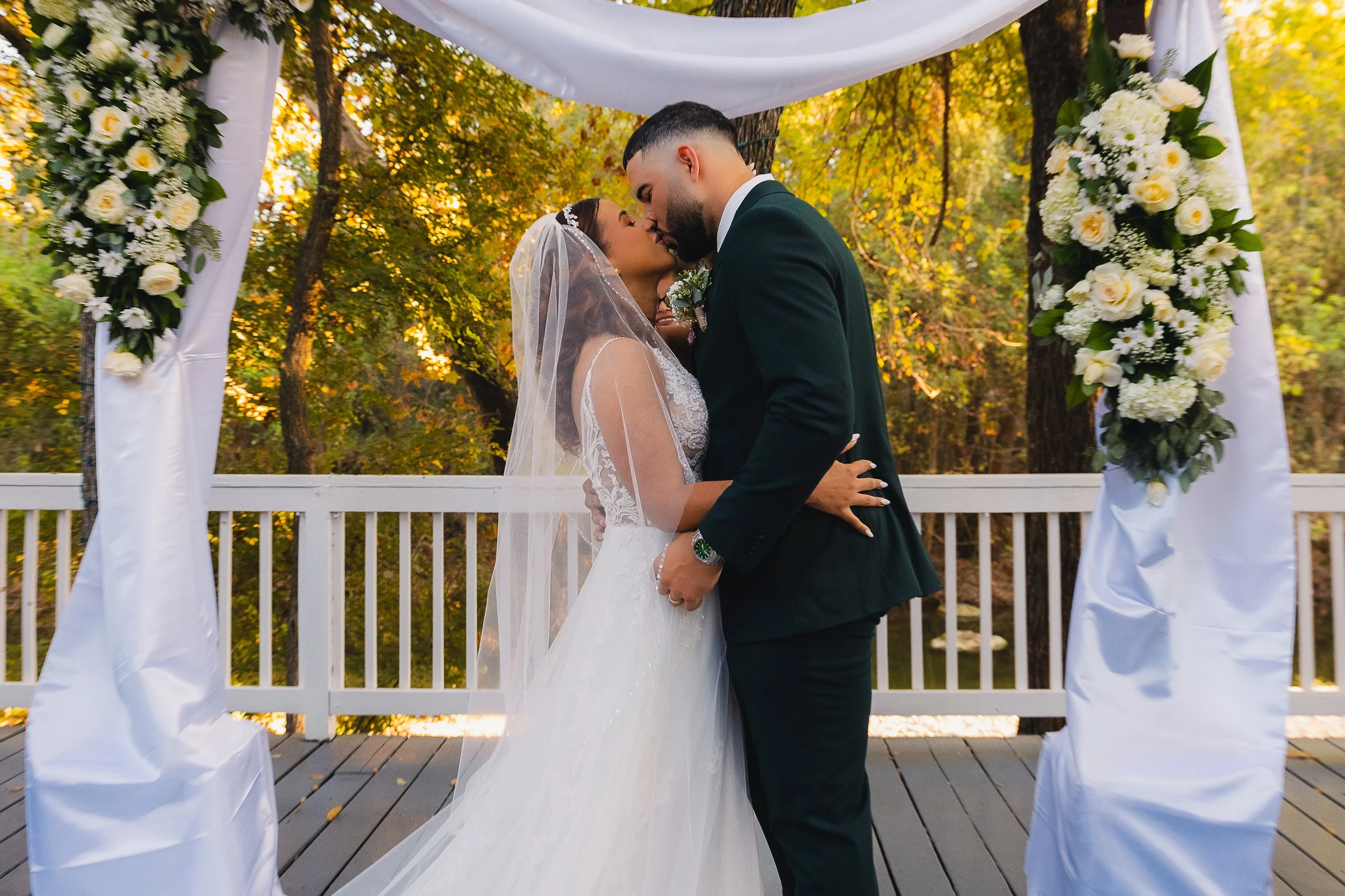 A bride and groom sharing a kiss under a floral wedding arch outdoors during sunset.