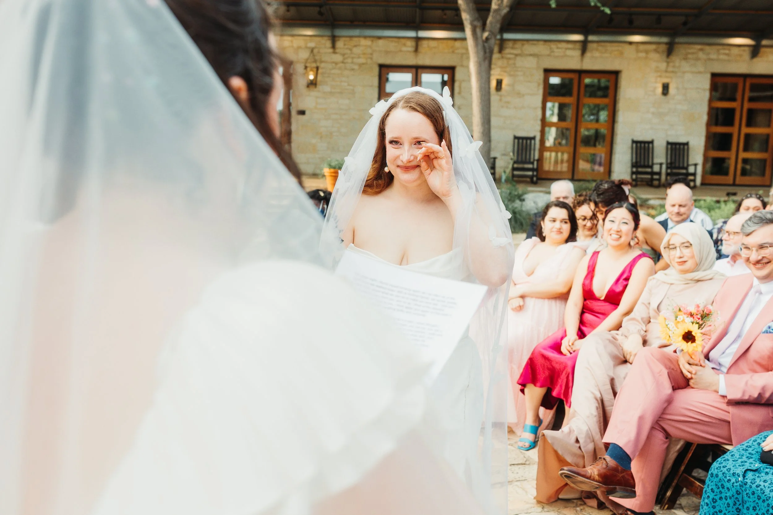 A bride with a veil wiping away tears during her wedding ceremony outdoors, with seated guests watching and smiling in the background.