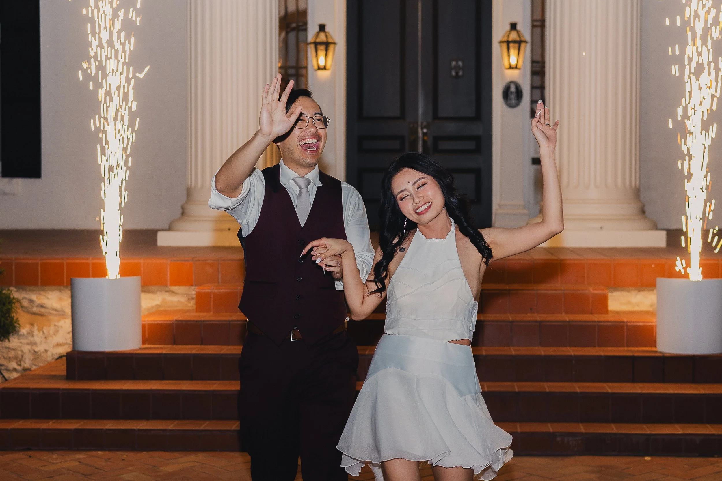 A couple dancing happily at an outdoor event with sparklers and a grand entrance in the background.