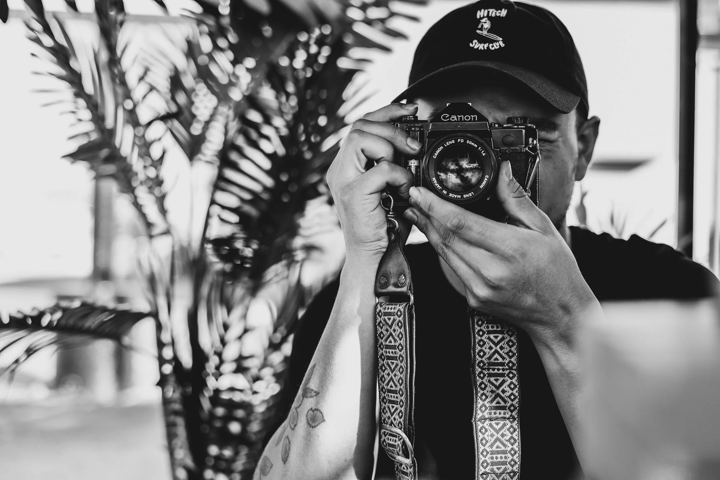A person taking a photo with a vintage Canon camera, wearing a dark cap and a patterned strap, with a large potted plant in the background.