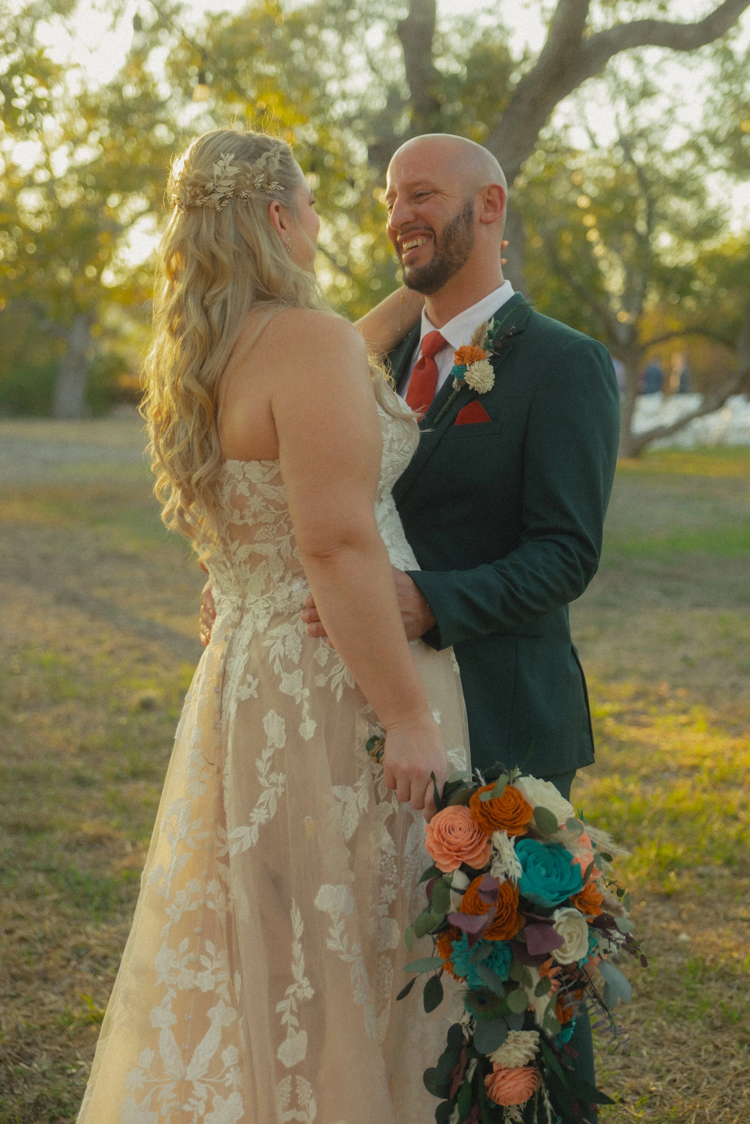 A bride and groom share a moment outdoors during sunset. The bride wears a lace wedding dress with floral embroidery and has long blonde hair with a hairpiece. The groom wears a dark suit with a white shirt, orange tie, and boutonniere. They smile at
