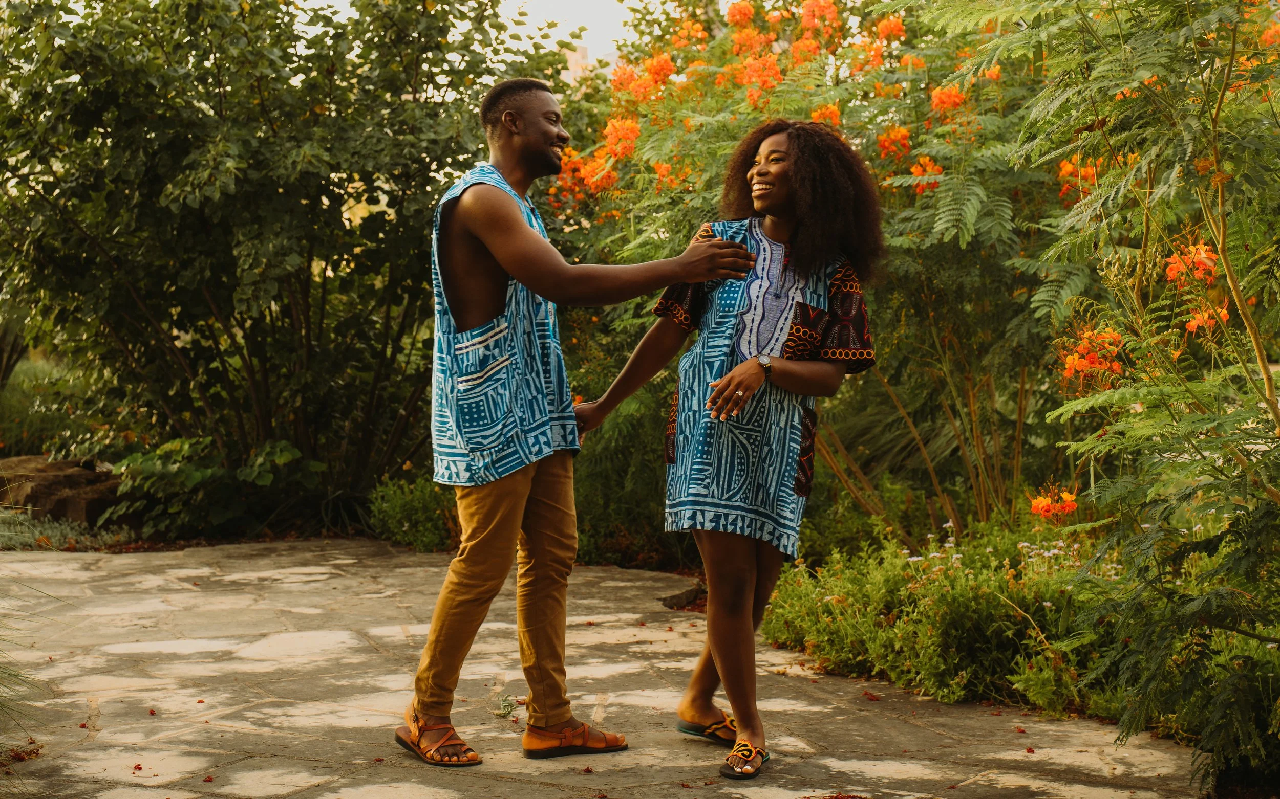 A man and woman holding hands and smiling while walking outdoors on a stone path, surrounded by lush greenery and orange flowers.
